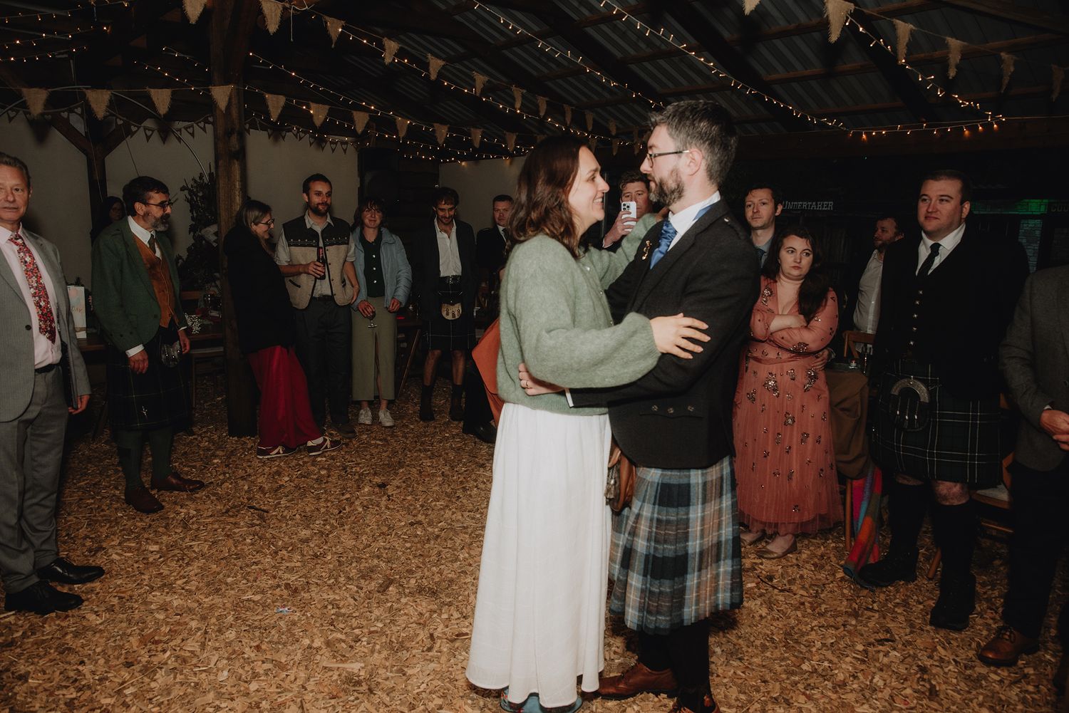 Newlywed couple dancing together in a decorated barn reception hall surrounded by smiling wedding guests.
