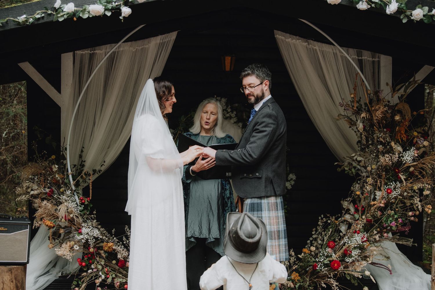 Bride and groom hold hands before officiant under circular arch adorned with flowers at forest wedding.