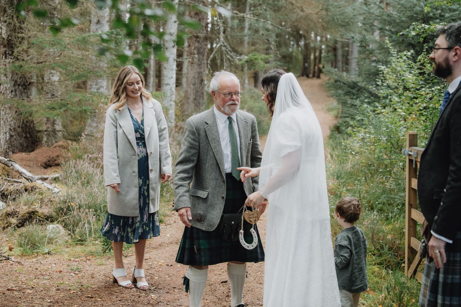 Bride in white veil walks woodland path with elderly man in tartan kilt and young child nearby.