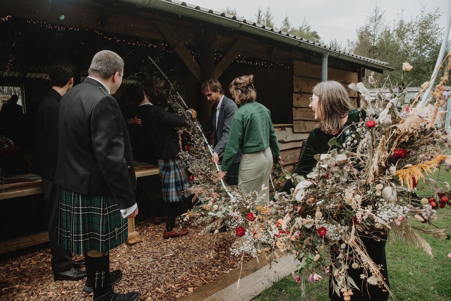 Wedding guests in kilts admire large dried floral arrangements outside a rustic barn.