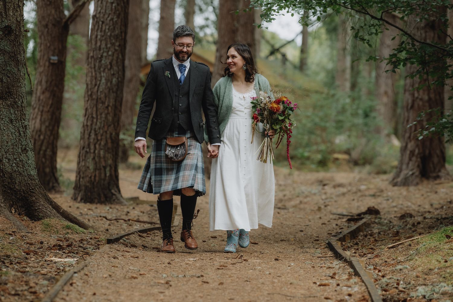 A bride and groom walk through a woodland forest, bride holding a colorful autumn bouquet.