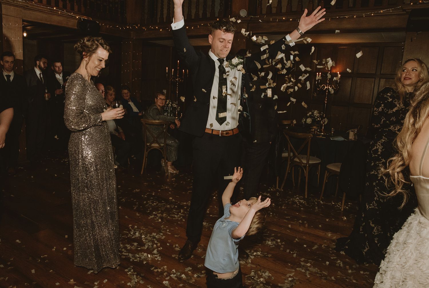 Joyful wedding celebration with confetti toss, groom in black suit, child playing, guests seated in warmly lit venue.