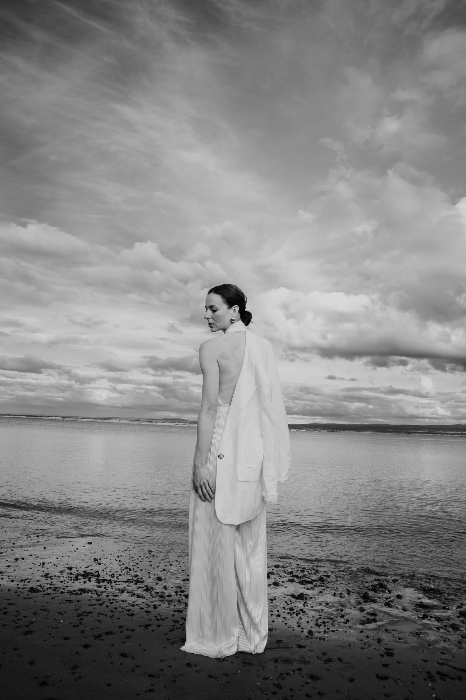 Black and white portrait of woman in white backless dress and blazer standing on beach with overcast sky.