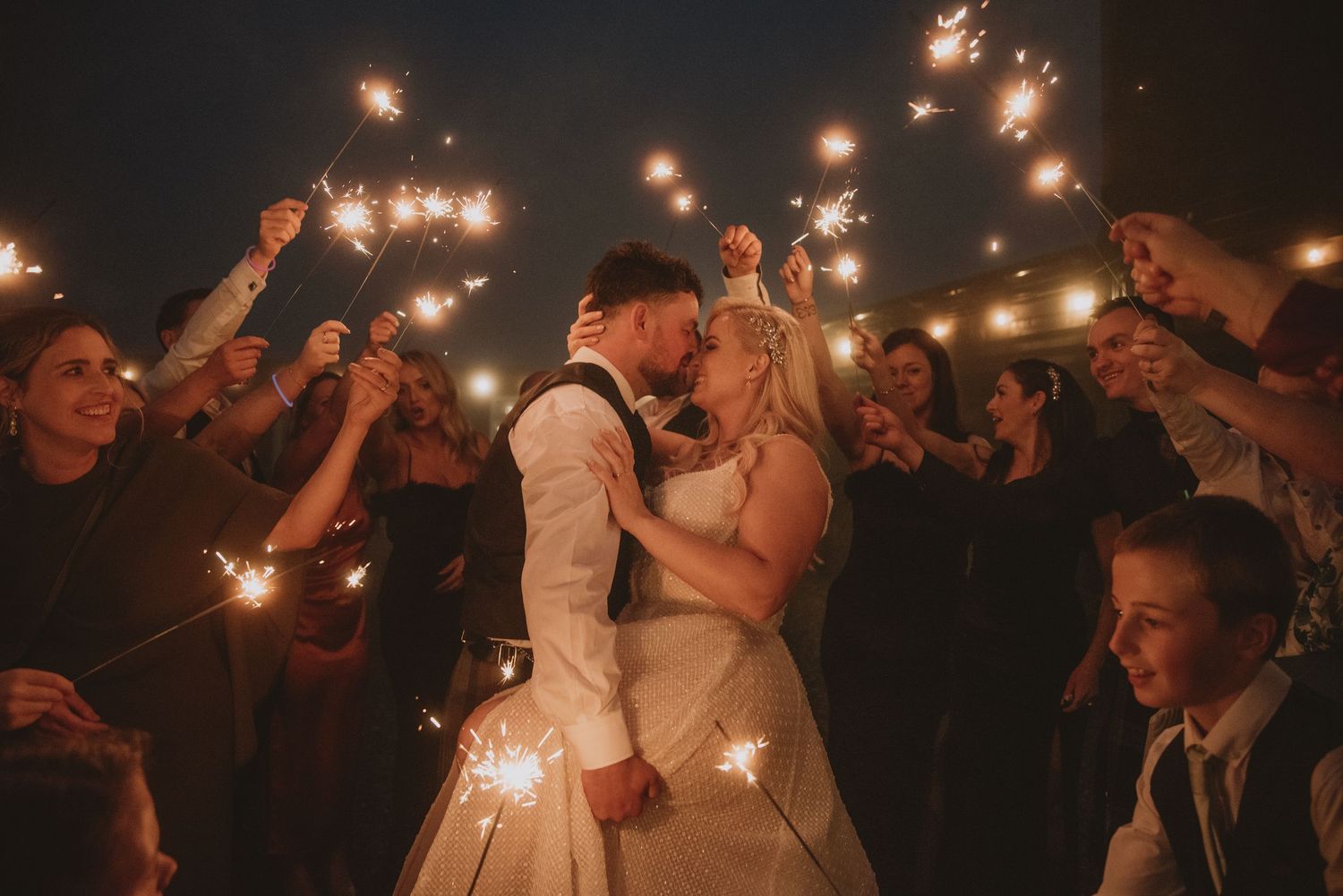 Bride and groom share a romantic kiss surrounded by guests holding sparkling sparklers at night.