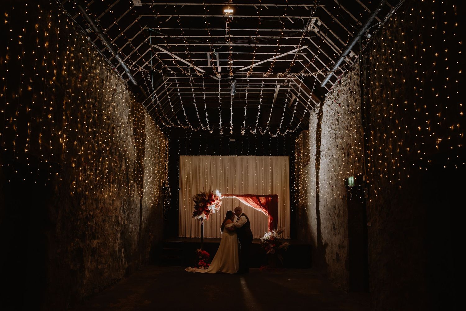 Bride and groom share romantic dance beneath stunning fairy light canopy in atmospheric barn wedding venue.