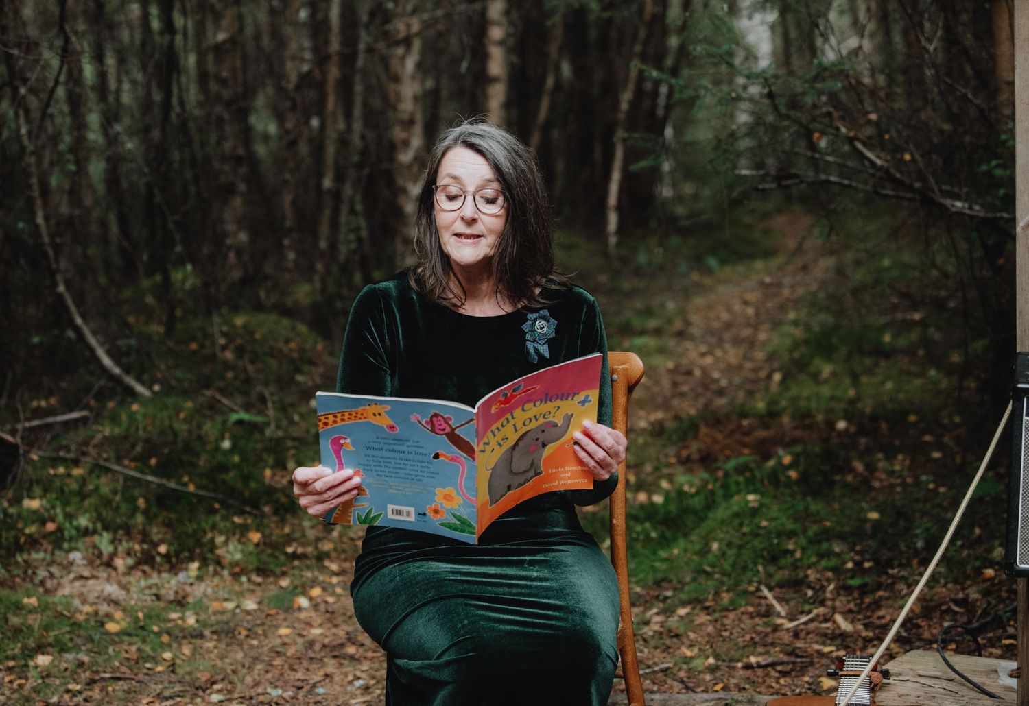 Woman sits on chair reading colorful childrens book in woodland forest setting.
