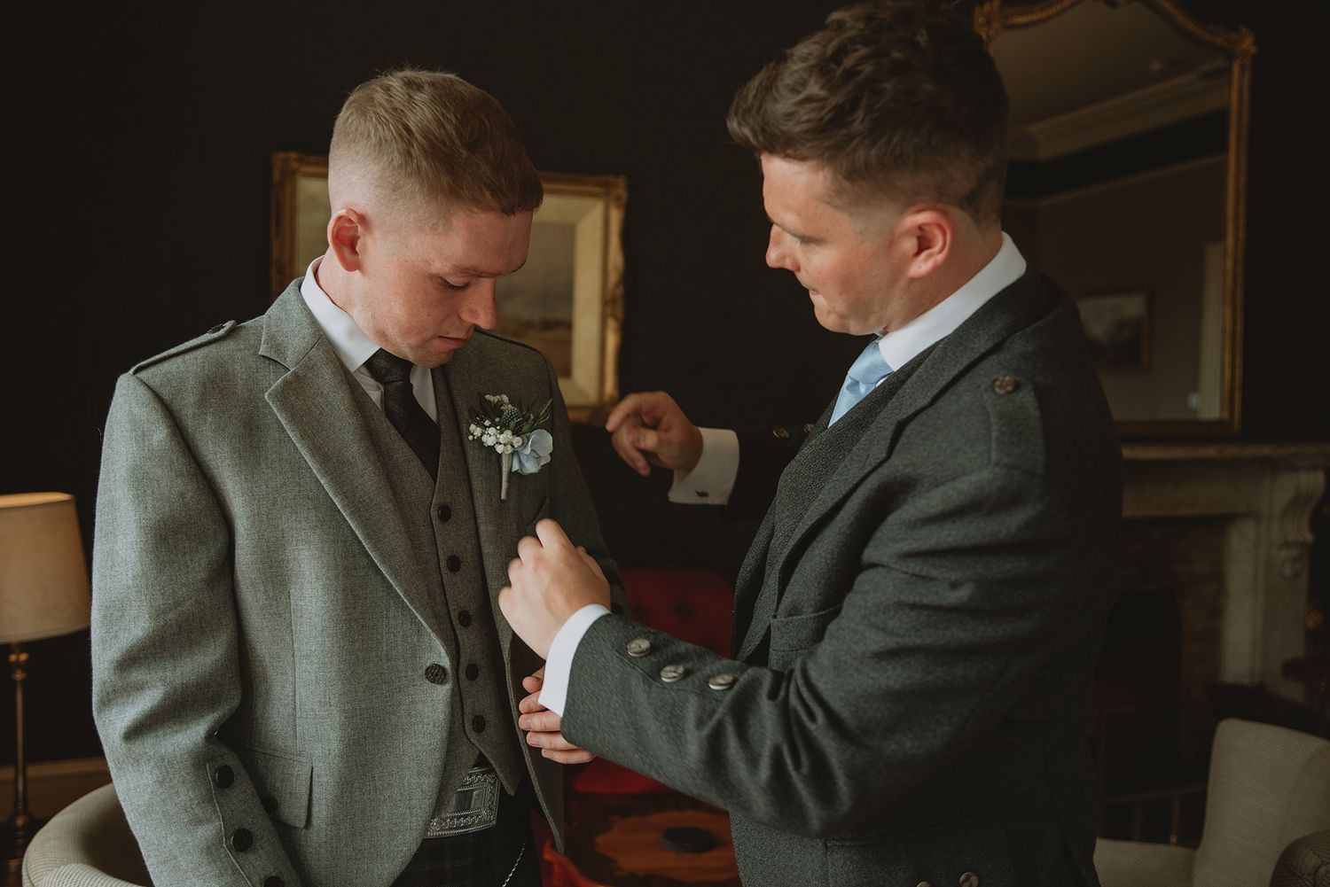 Groomsman helping groom with boutonniere on grey tweed suit before Scottish wedding ceremony.