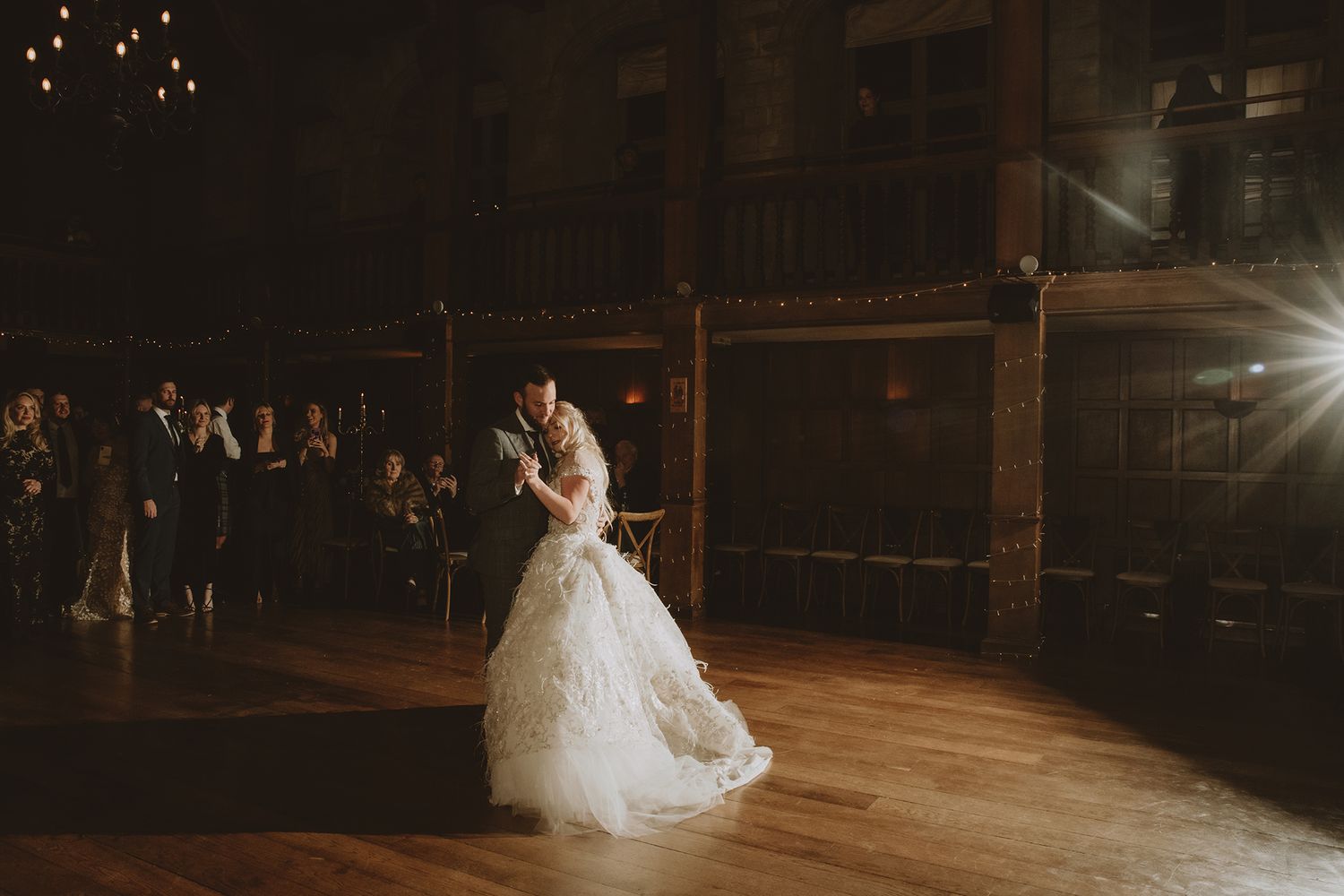 Bride and groom share their first dance on a wooden floor in a dimly lit rustic wedding venue with guests watching.