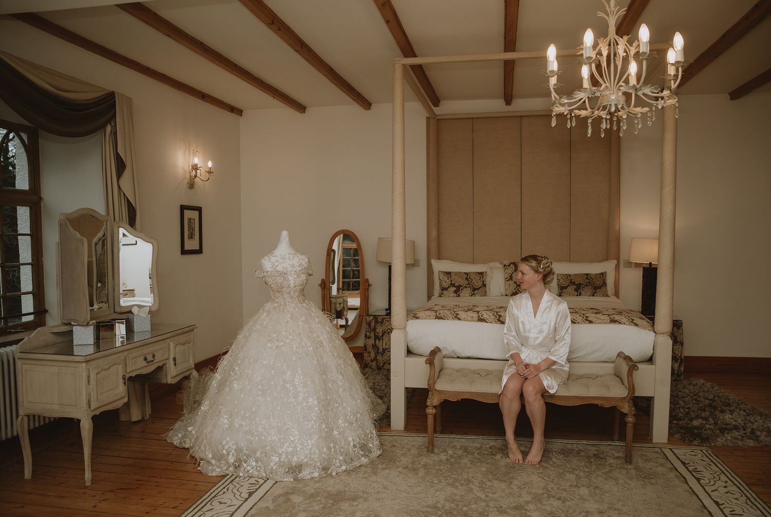 Bride in white robe sits on bench beside elegant wedding gown displayed on mannequin in luxurious bridal suite.