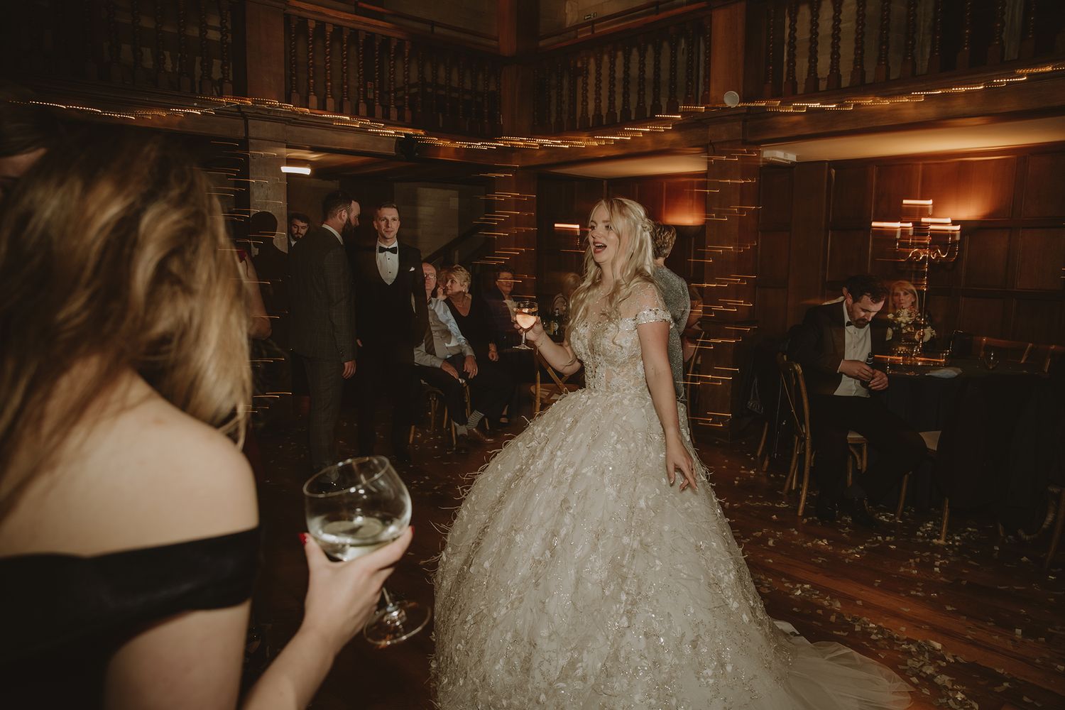 Bride in a stunning lace wedding gown holds a champagne glass while dancing at an elegant rustic wedding reception.