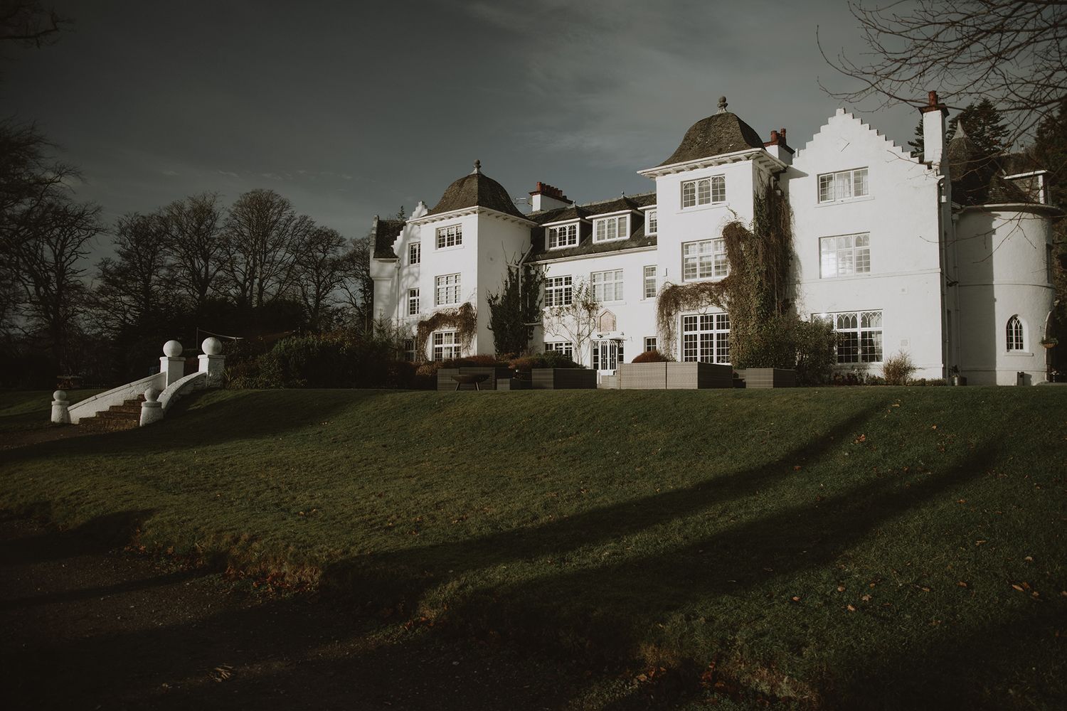 A grand white manor house with ivy-covered walls set on lush green grounds under a dramatic cloudy sky.