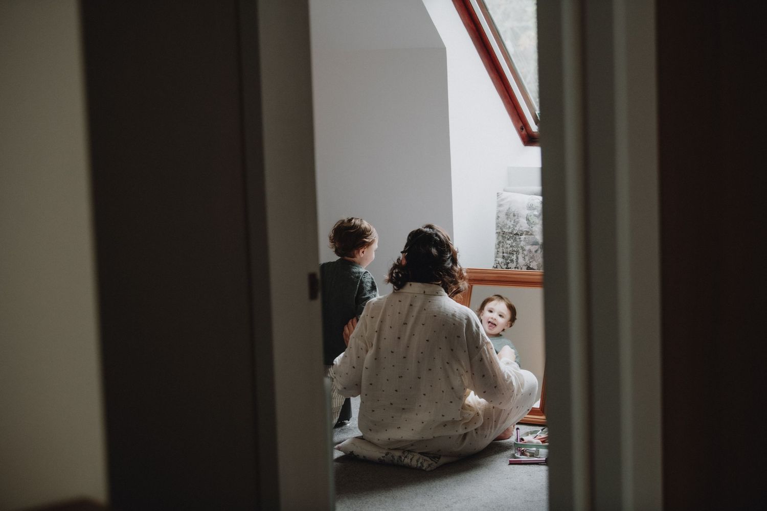 Bride in white robe sits on floor with two children in a bright attic room on wedding day.