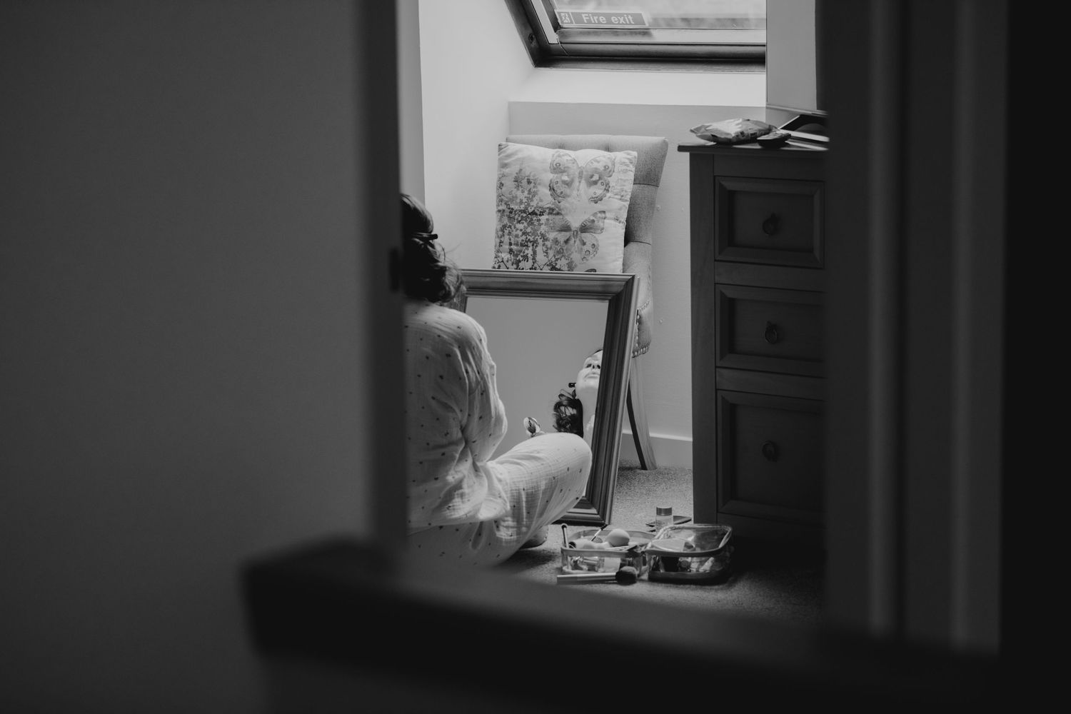 Black and white photo of a bride sitting on the floor getting ready in a sunlit attic bedroom.