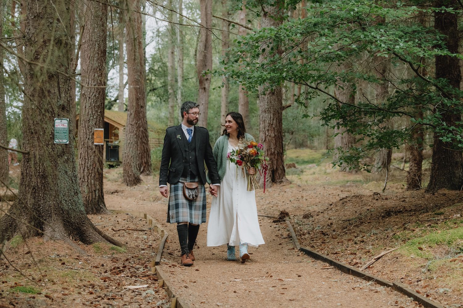 Newlywed couple strolling along a woodland path, groom in tartan kilt holding bride in white dress.