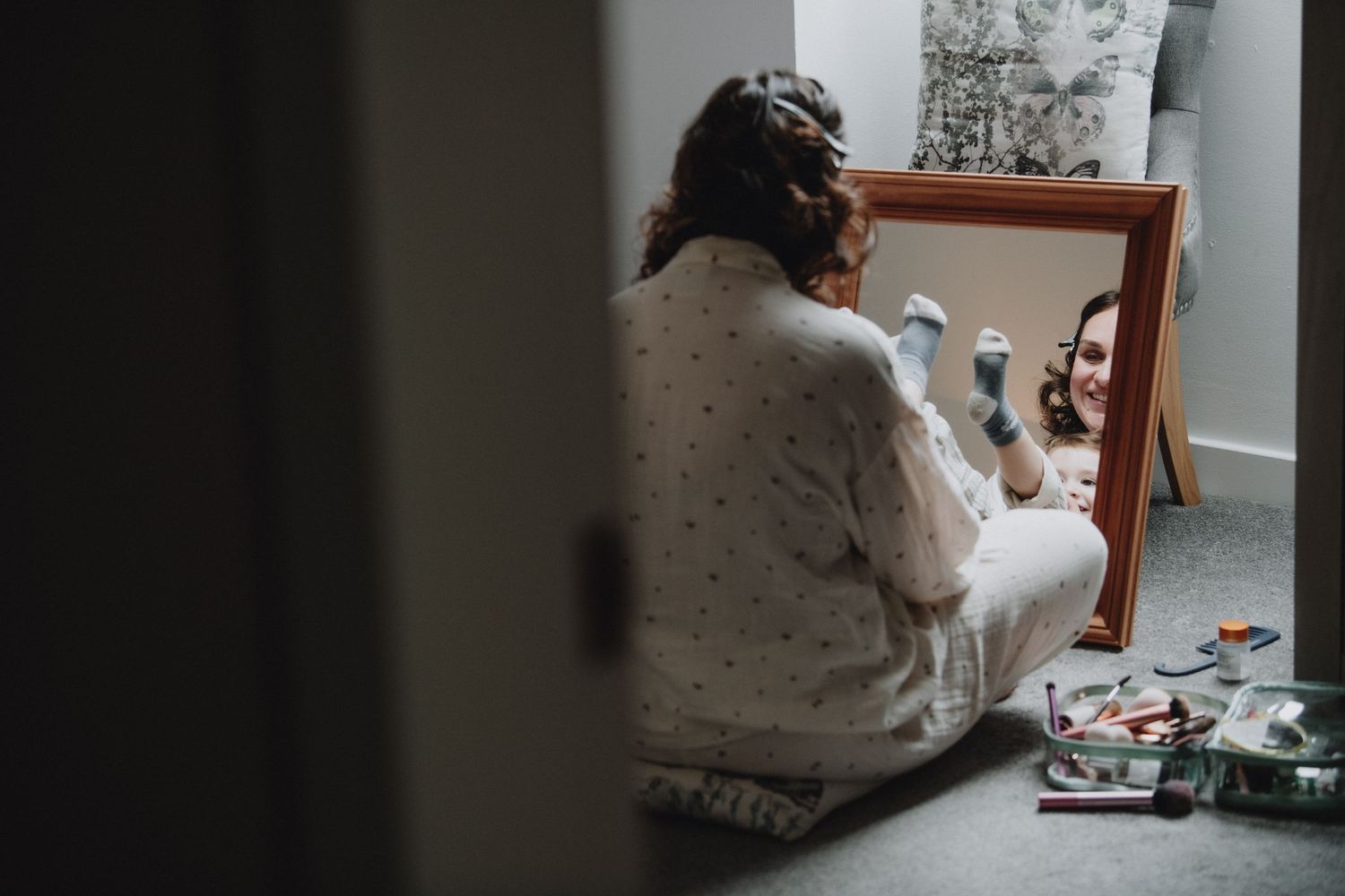 Bride in polka dot robe applies makeup seated before a mirror with wedding accessories nearby.