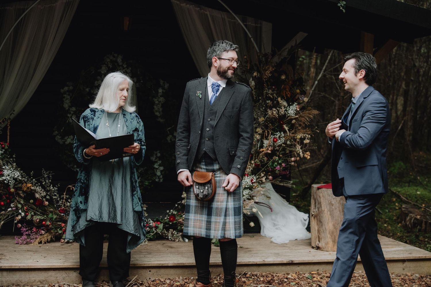 Groom in tartan kilt laughs with best man during outdoor Scottish woodland wedding ceremony.
