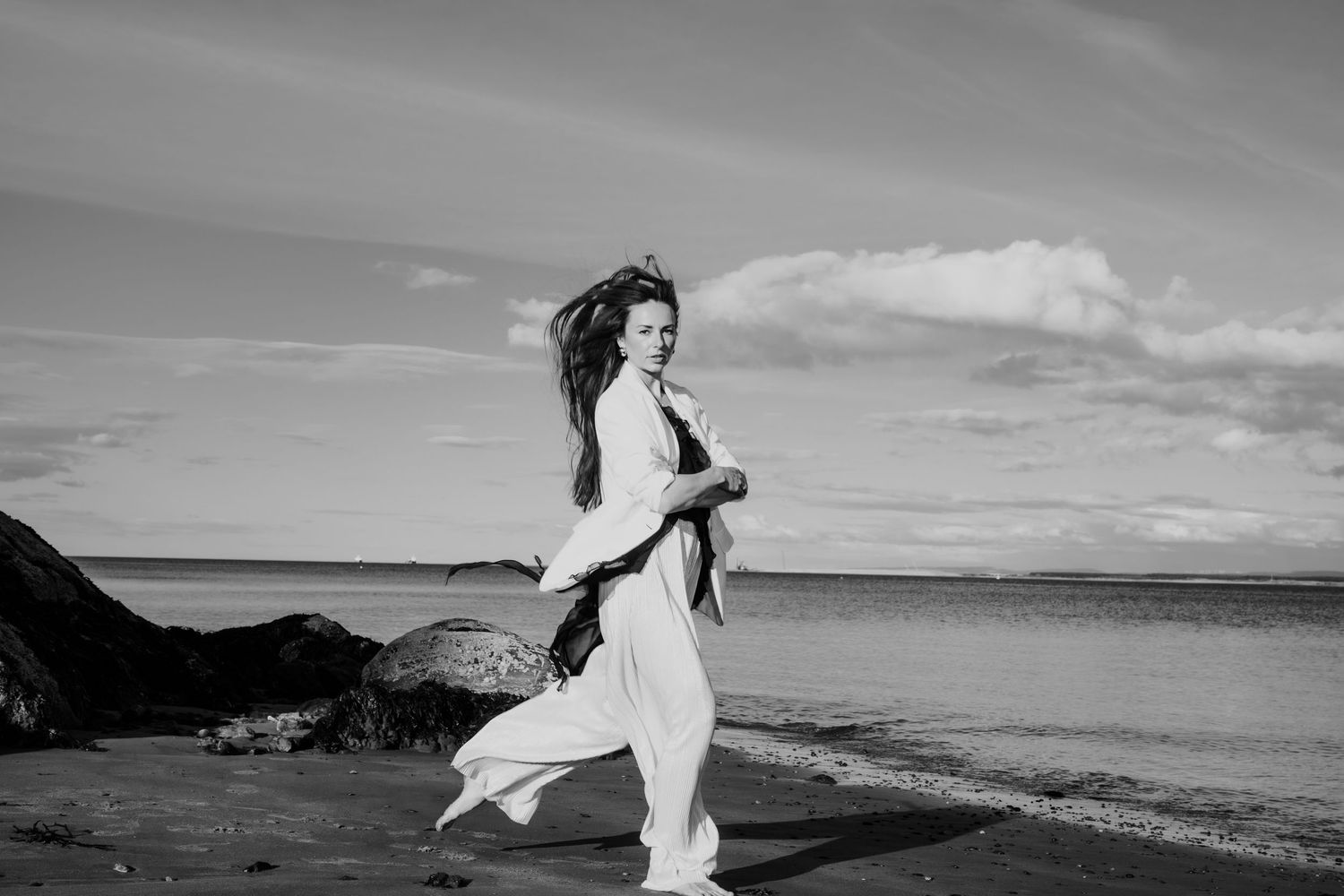 Black and white photo of a woman in white outfit walking along a rocky seaside beach.