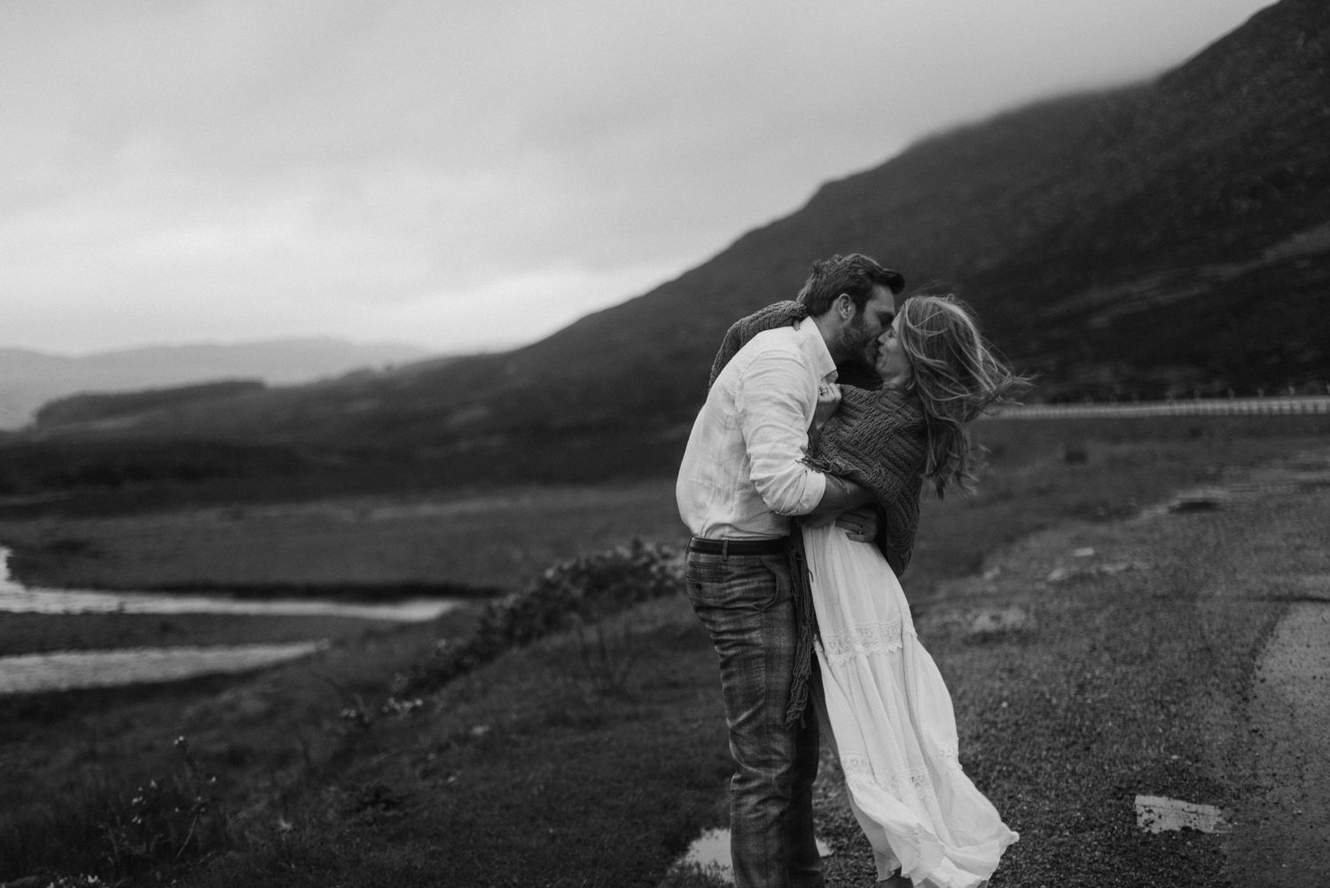 Black and white photo of couple kissing on rural Scottish Highland road with mountain backdrop.
