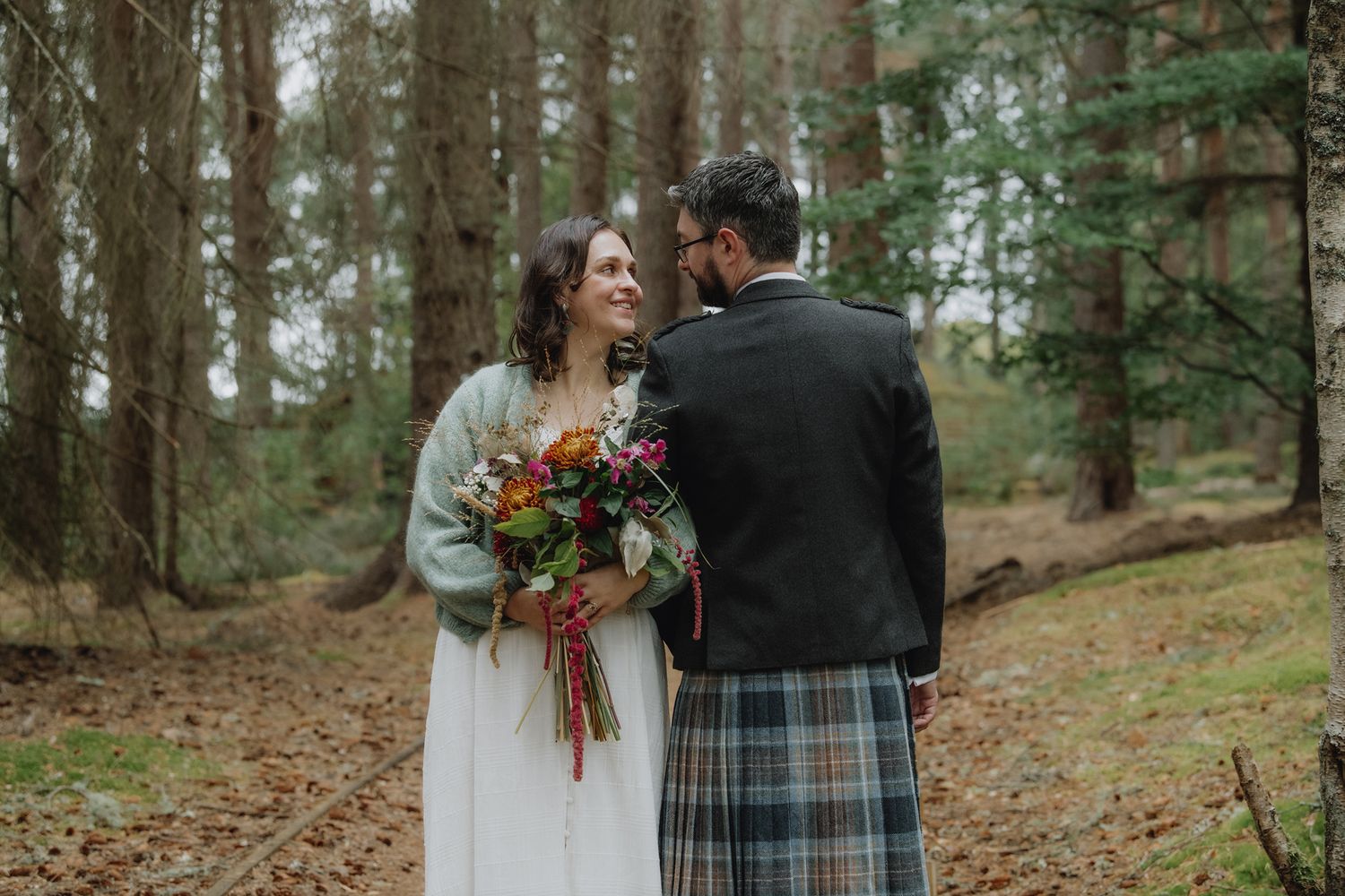 Bride and groom sharing a tender moment on a forest path, groom in tartan kilt holding colorful bouquet.