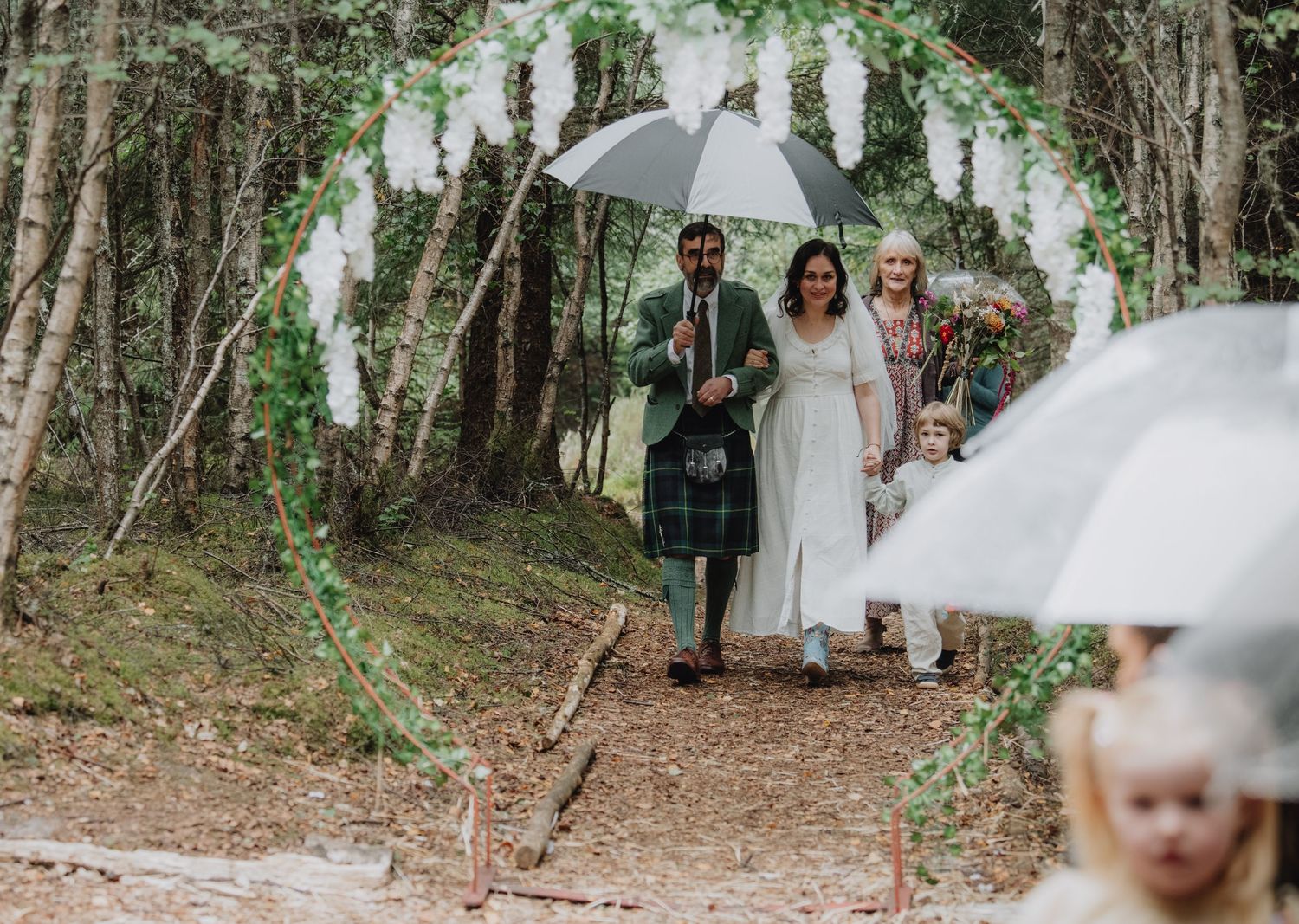 Bride and groom walk through woodland wedding arch with umbrellas in Scottish forest ceremony.