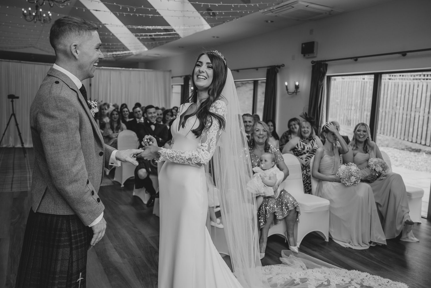 Black and white wedding ceremony photo of bride in veil and groom in tartan kilt exchanging vows before seated guests.