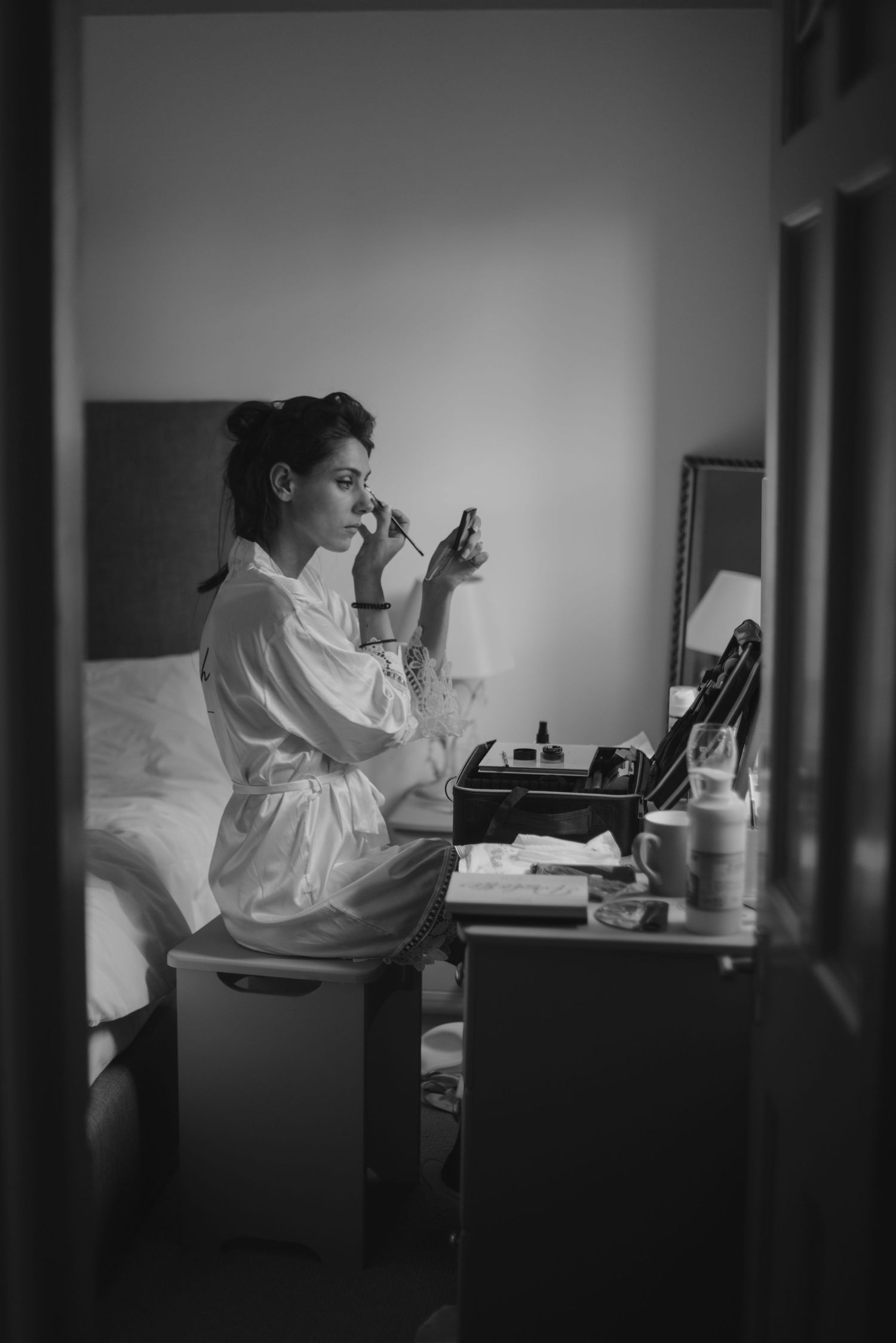 Black and white photo of a bride in a white robe applying makeup at a vanity table in a hotel room.