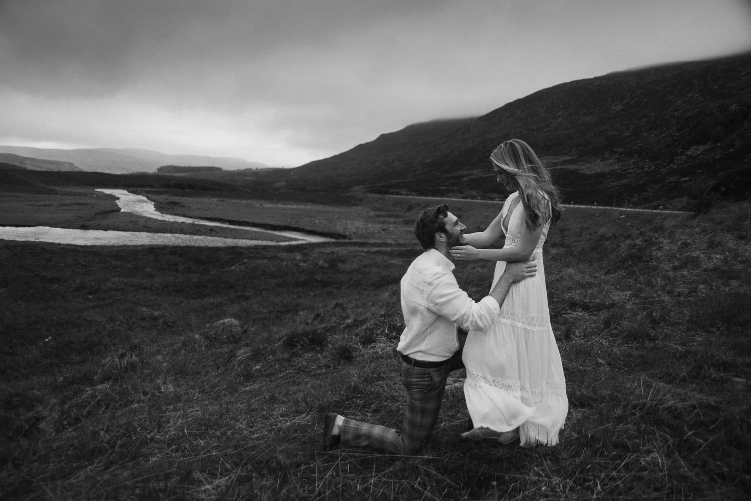 Groom on one knee holding brides hands in dramatic black and white Scottish Highland landscape.