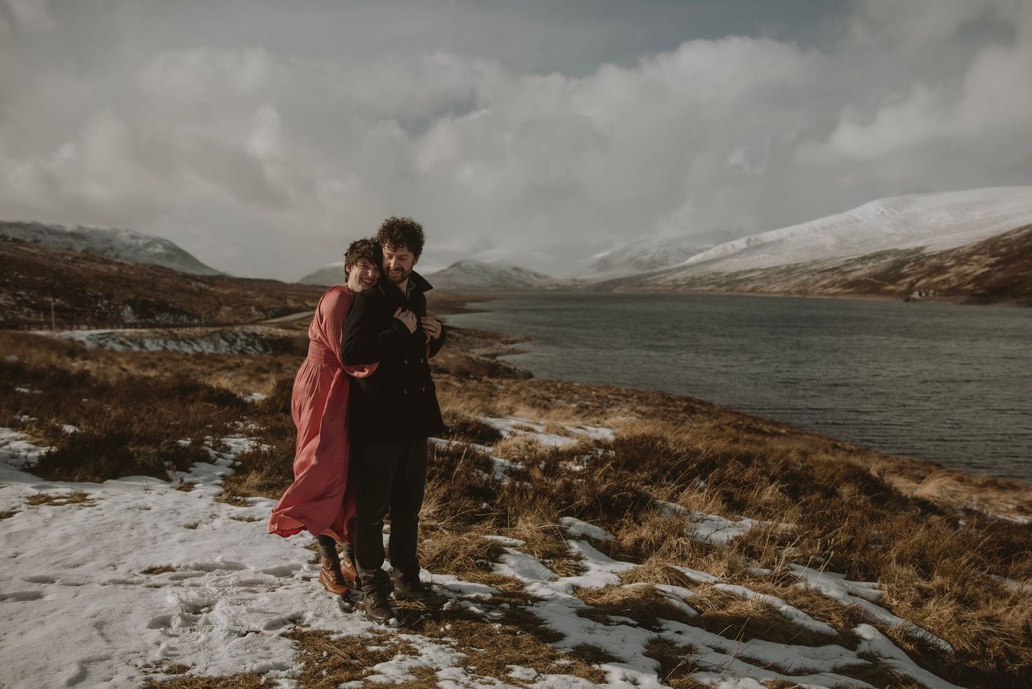 Couple embracing by a snowy loch in the Scottish Highlands, surrounded by winter mountains and dramatic cloudy skies.