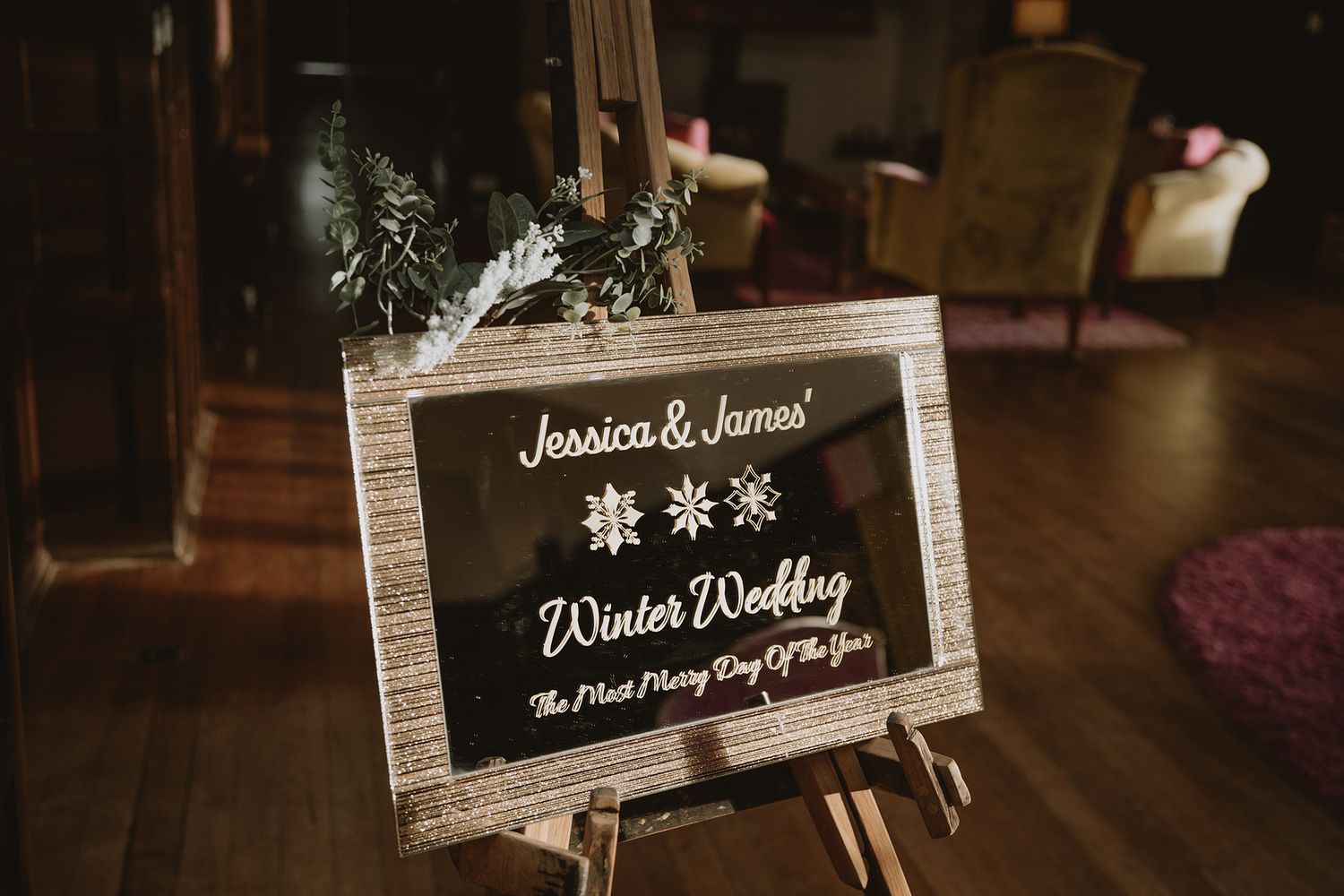 A rustic winter wedding welcome sign for Jessica and James displayed on a wooden easel with snowflake decorations and greenery.