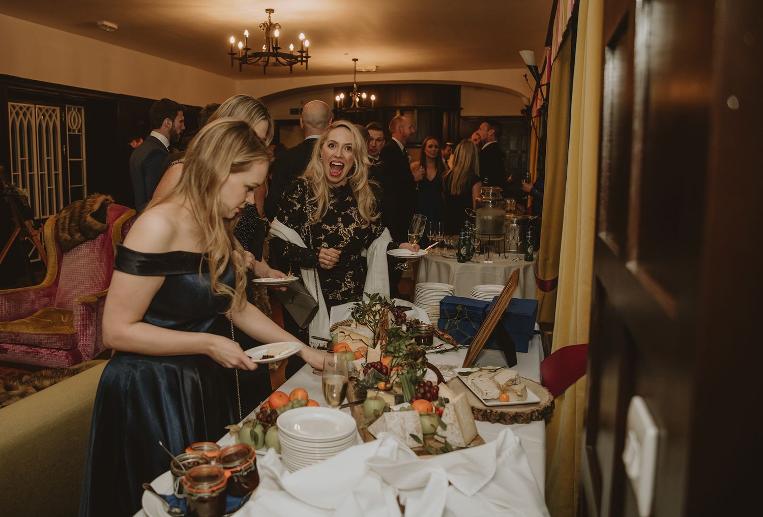 Guests serving themselves at an elegant dinner party buffet table with floral centerpieces in a warmly lit venue.
