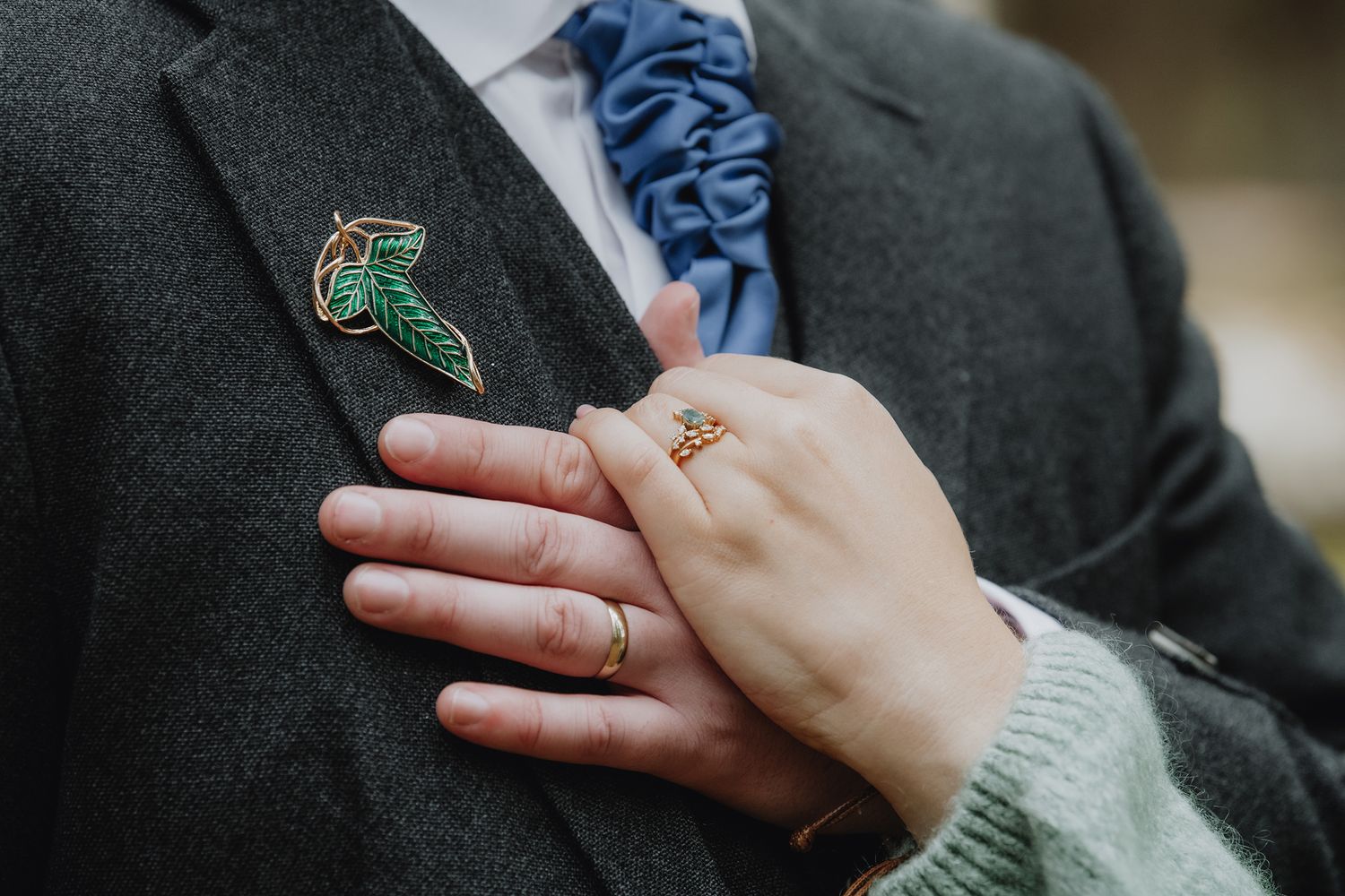 Close-up of bride's hand on groom's grey tweed jacket featuring a decorative leaf brooch and blue tie.
