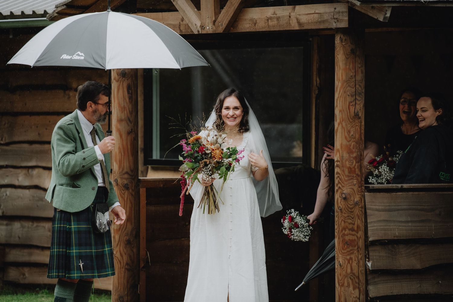 Radiant bride in white veil holding wildflower bouquet sheltered by umbrella outside a rustic log cabin.