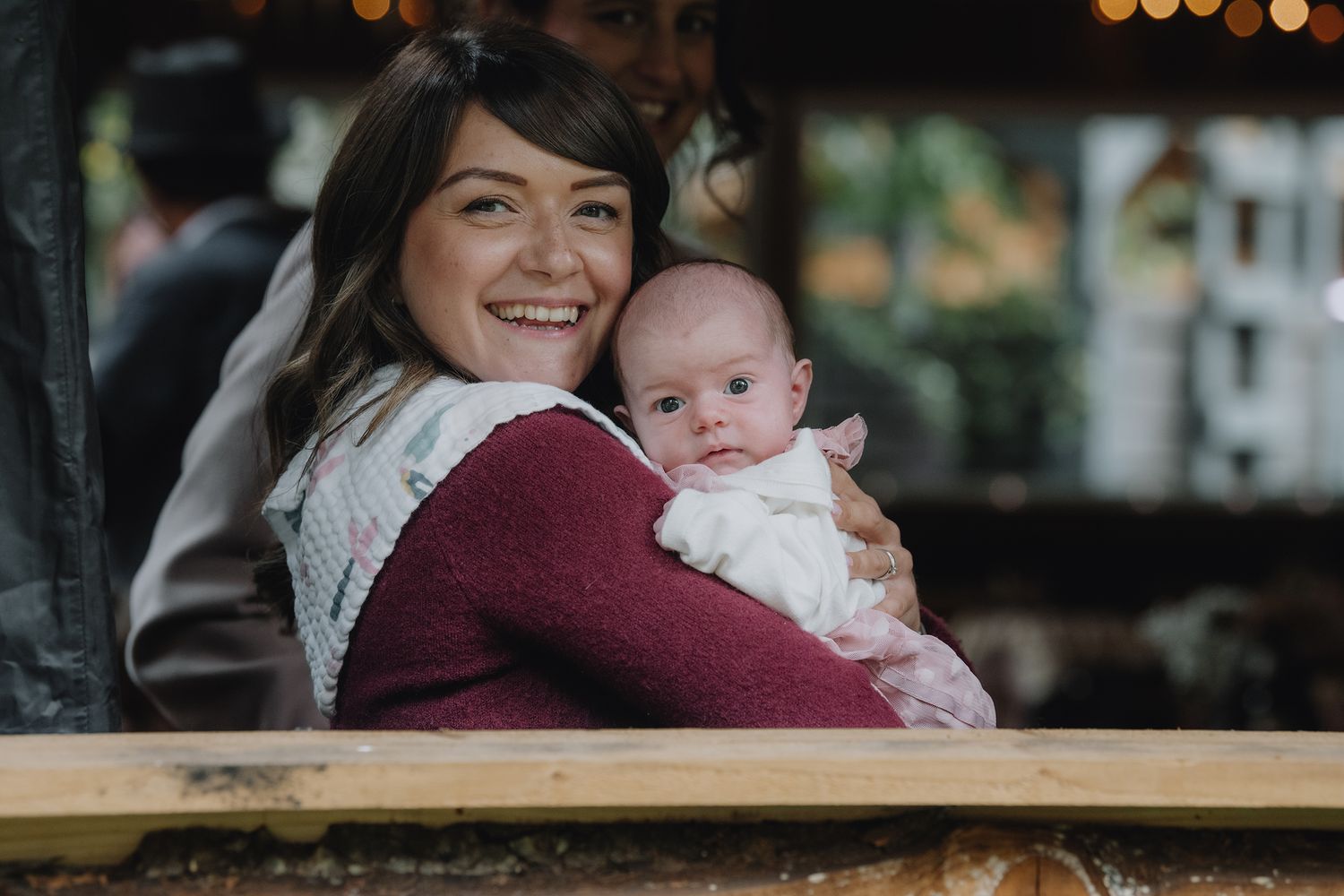 A smiling dark-haired woman holds a wide-eyed baby at an outdoor event.