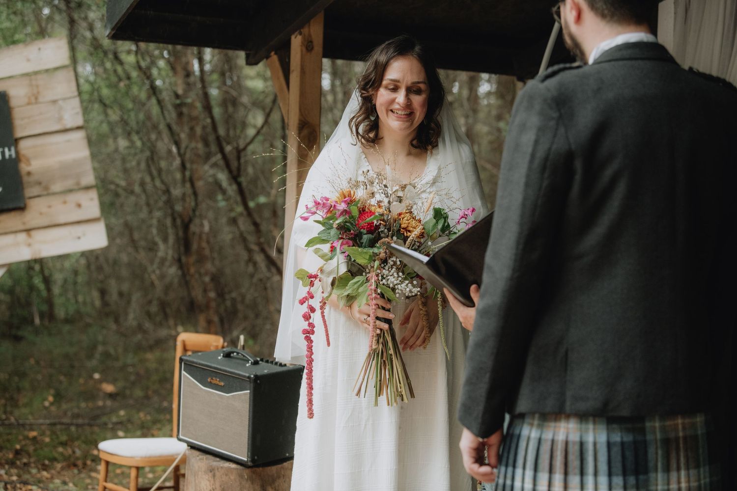 Smiling bride holds colorful wildflower bouquet facing groom in tartan kilt at woodland wedding.