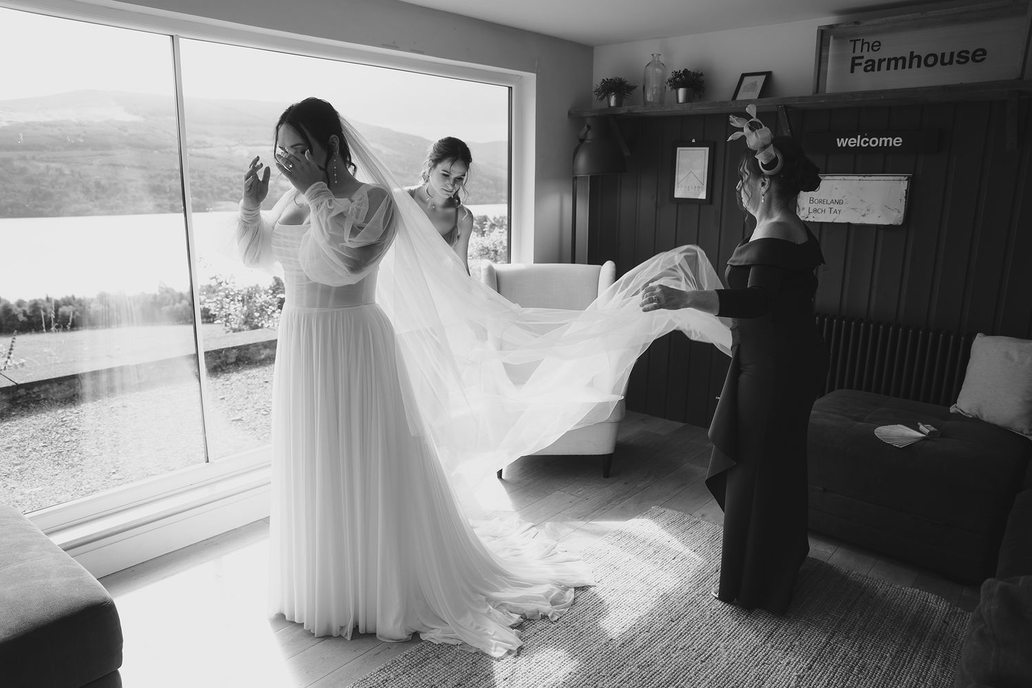 Black and white photo of bride being helped into wedding dress by bridesmaids with scenic lake view.