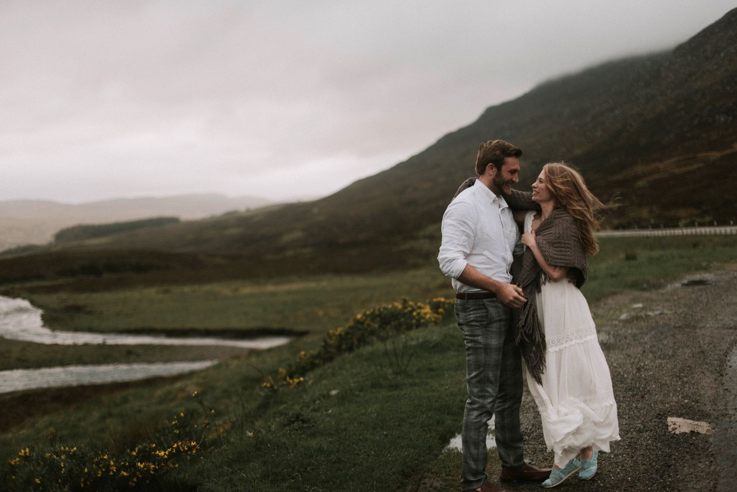 Couple sharing romantic moment on misty Scottish Highland road with green hills and river.