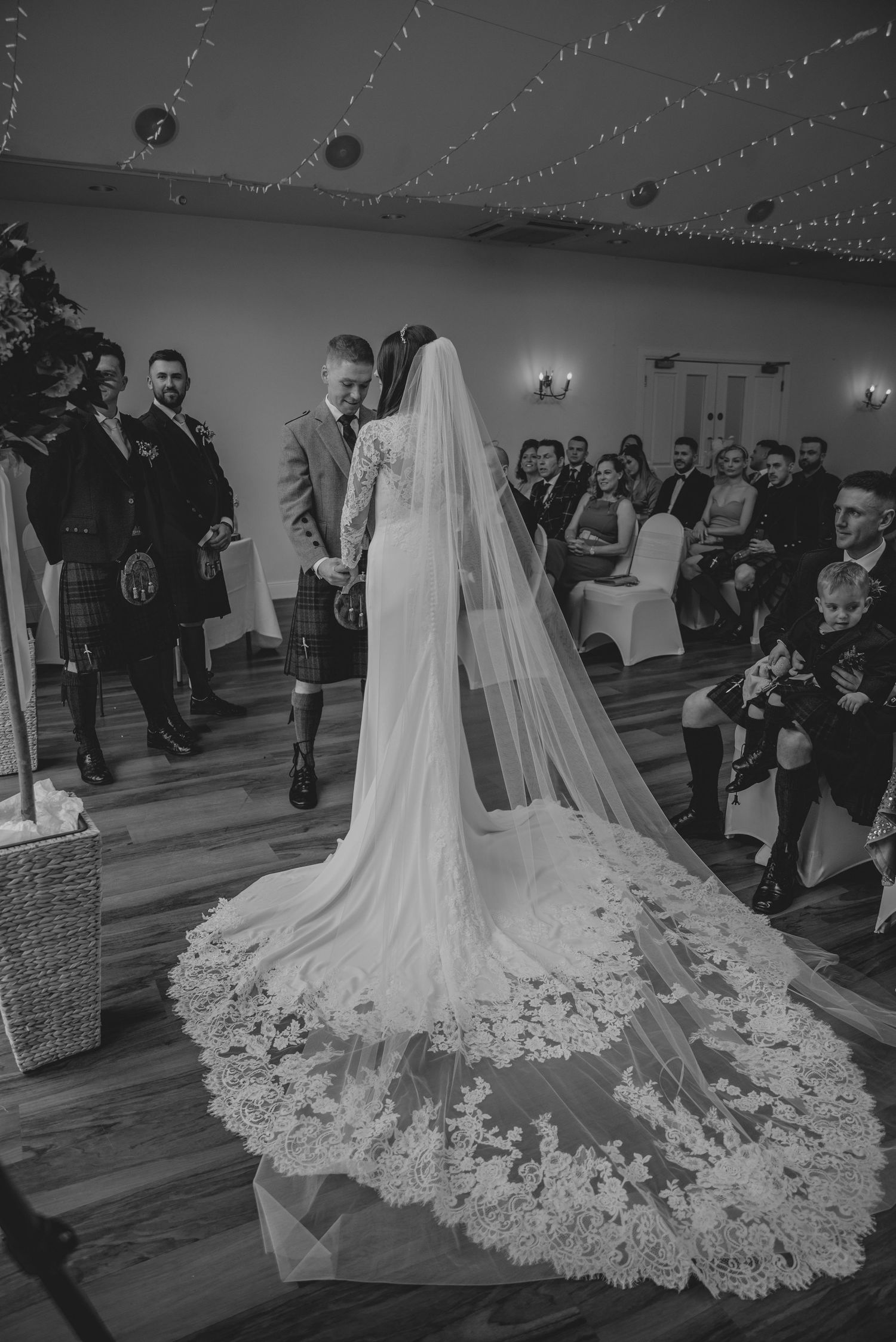 Black and white wedding ceremony photo showing bride in lace gown with cathedral veil, groom in kilt, guests seated in decorated venue.
