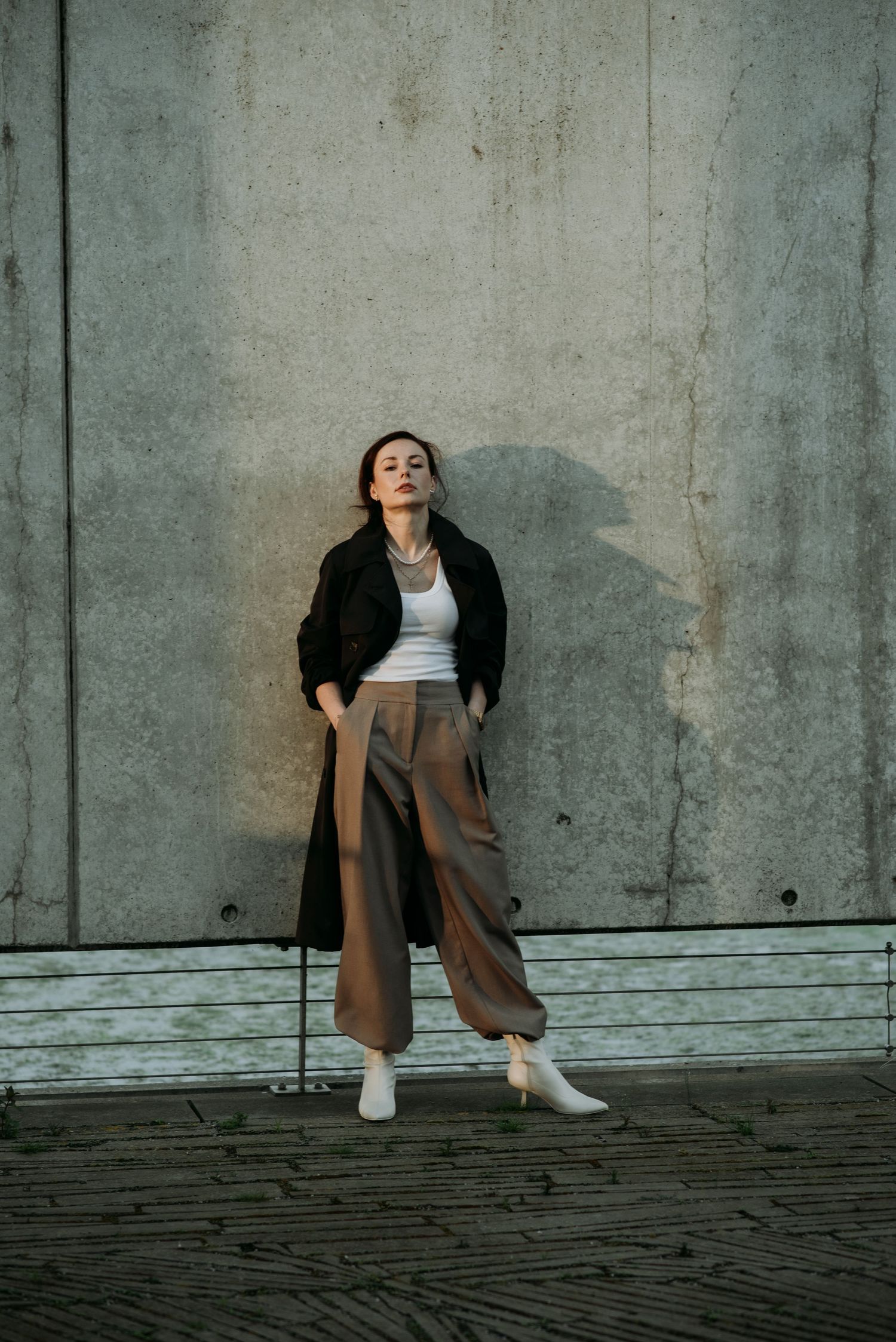 Woman in white top, brown wide-leg pants and black coat leans against concrete wall outdoors.