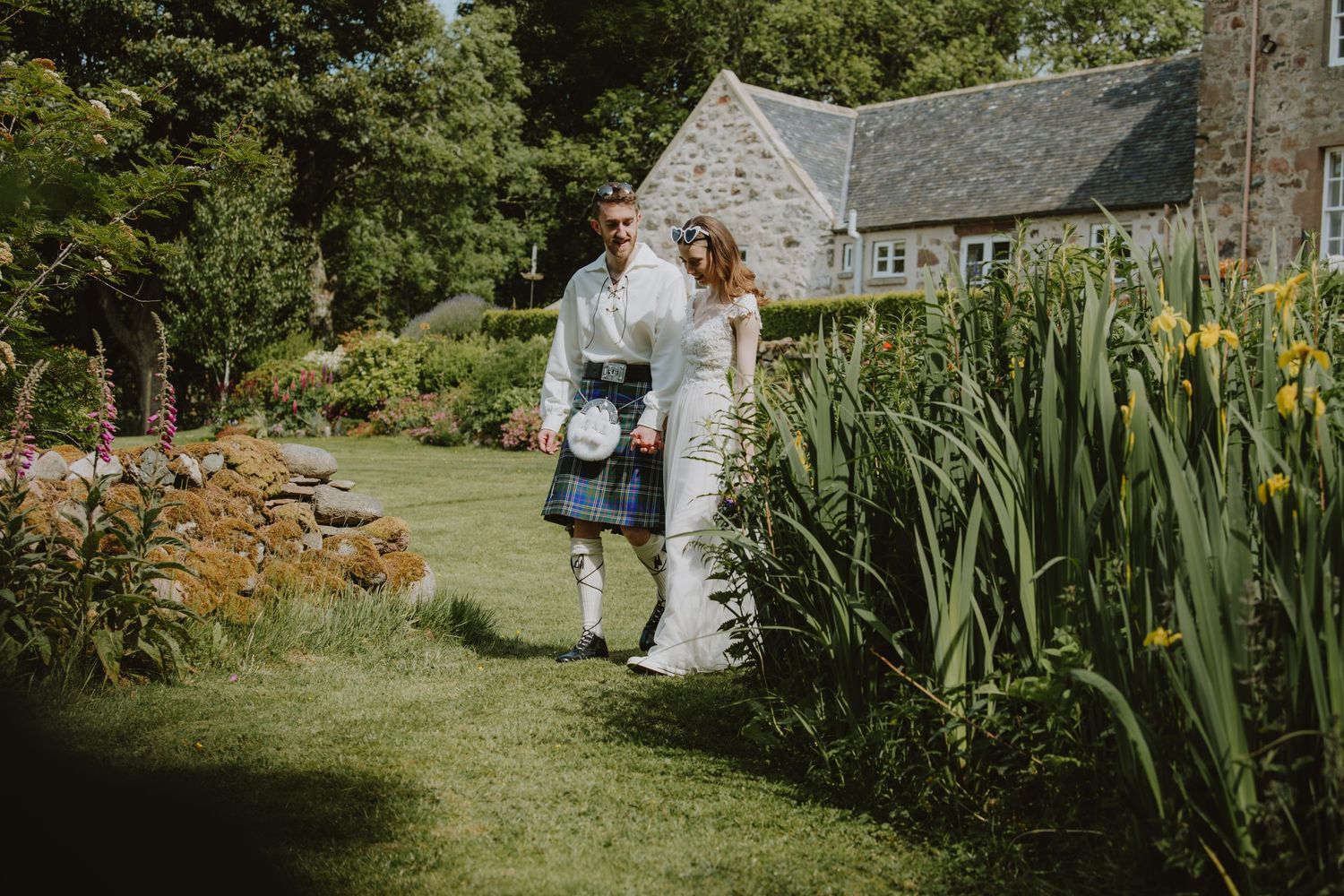 Bride and groom walking through lush garden, groom wearing traditional Scottish kilt, stone cottage in background.