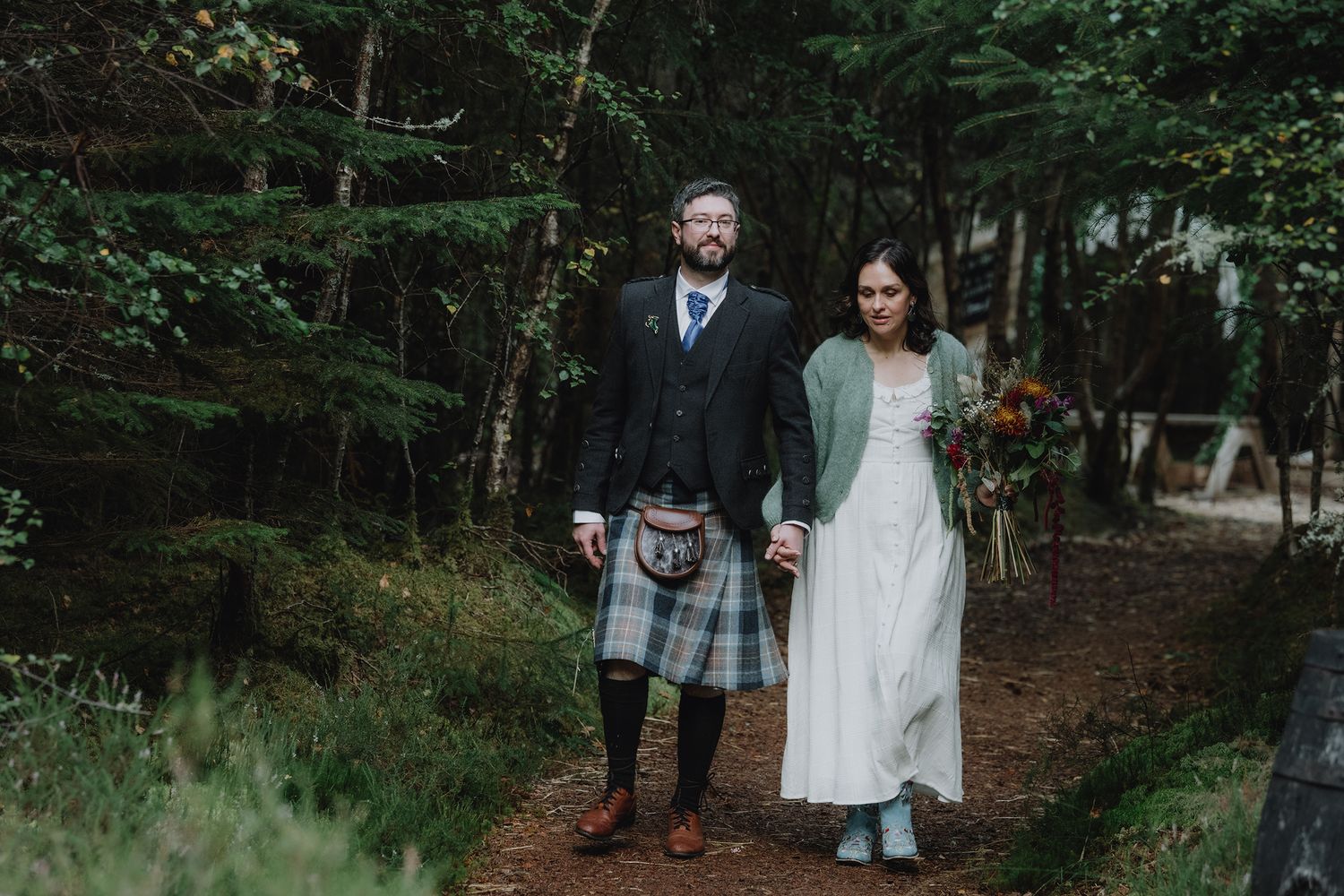 A bride and groom stand together on a forest path, groom dressed in traditional Scottish tartan kilt.