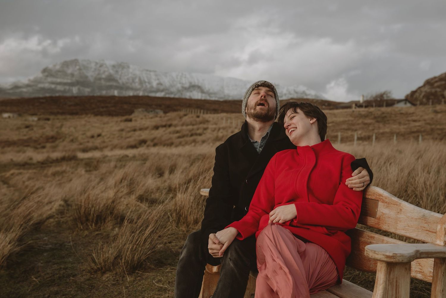 Couple sitting on wooden bench laughing in rustic field with snow-capped mountains, wearing red and black outfits.