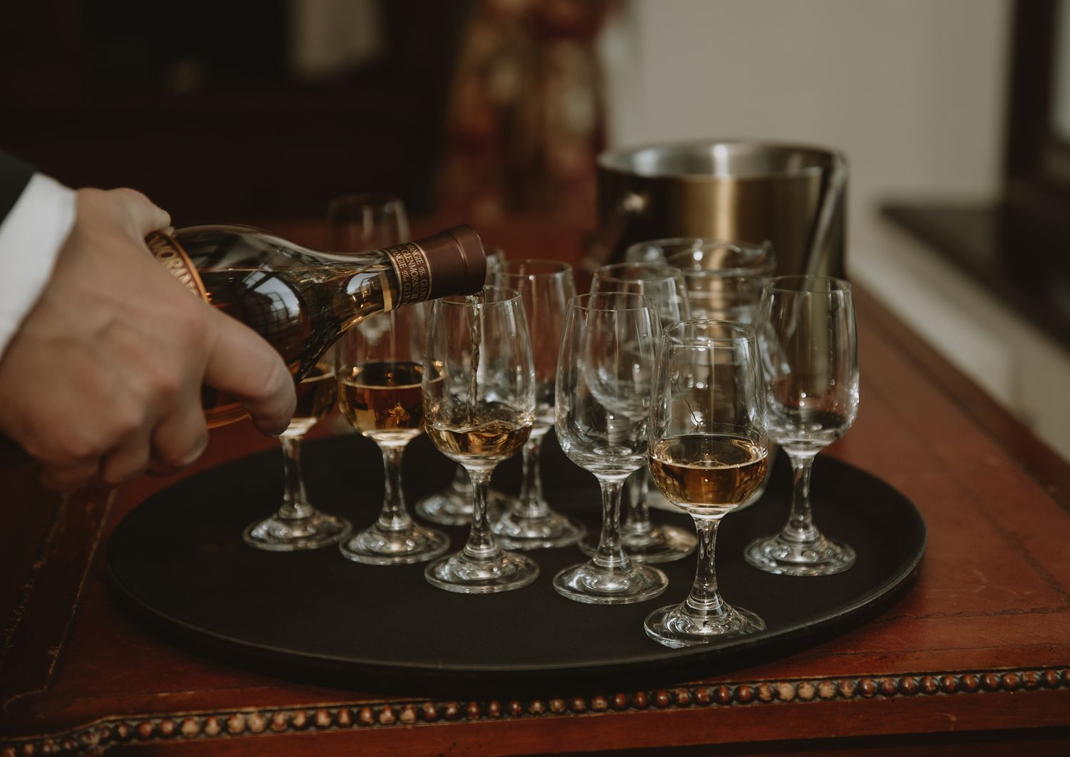 A bartender pouring amber whiskey into elegant crystal glasses arranged on a round black tray at a tasting event.