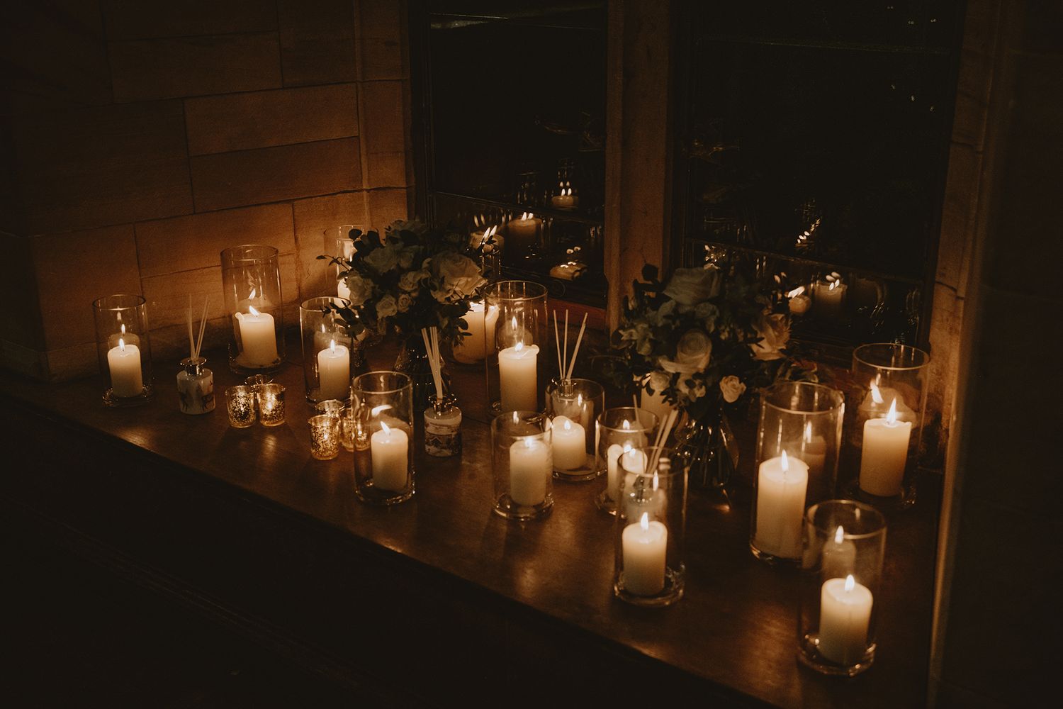 Romantic candlelit display with white pillar candles and floral arrangements on a wooden surface in dim lighting.