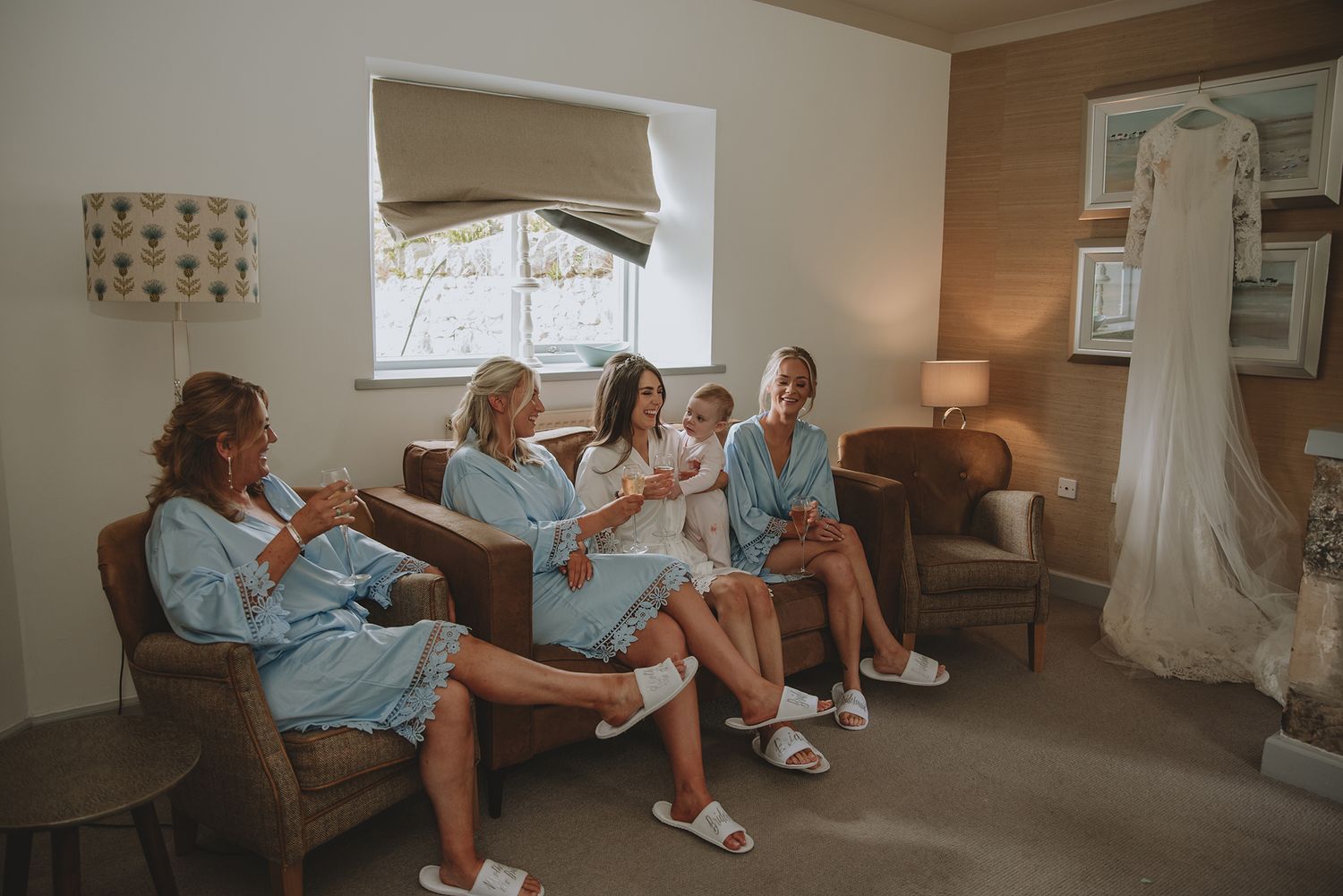 Bridesmaids in blue robes and white slippers relax together in a cozy room with a wedding dress hanging nearby.