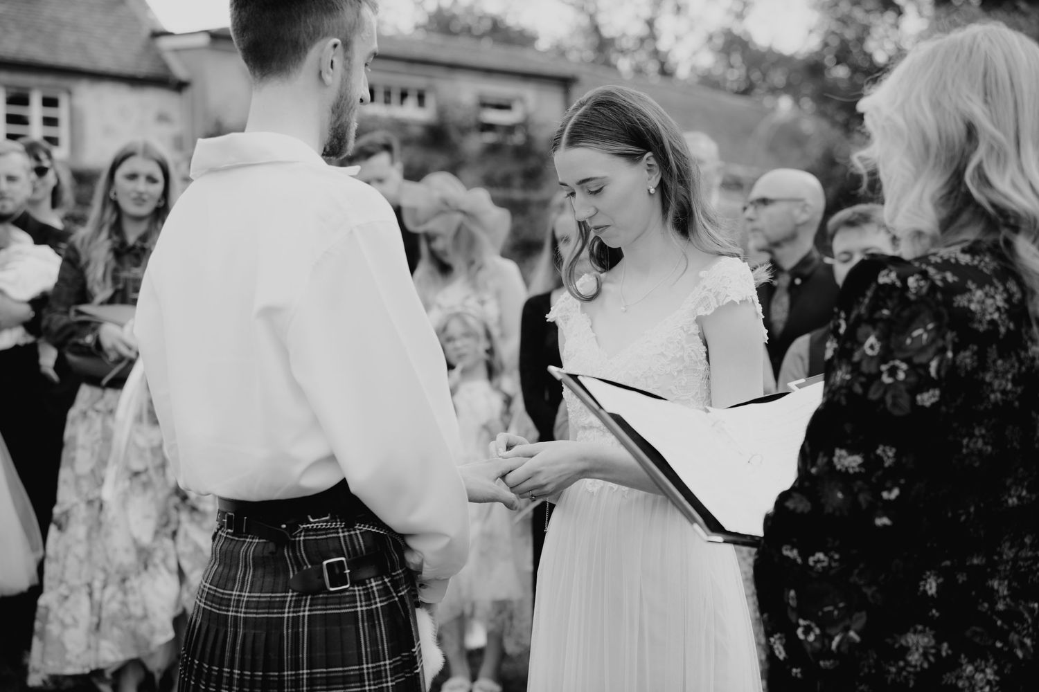 Bride places wedding ring on groom wearing a kilt during an outdoor ceremony, black and white photo.