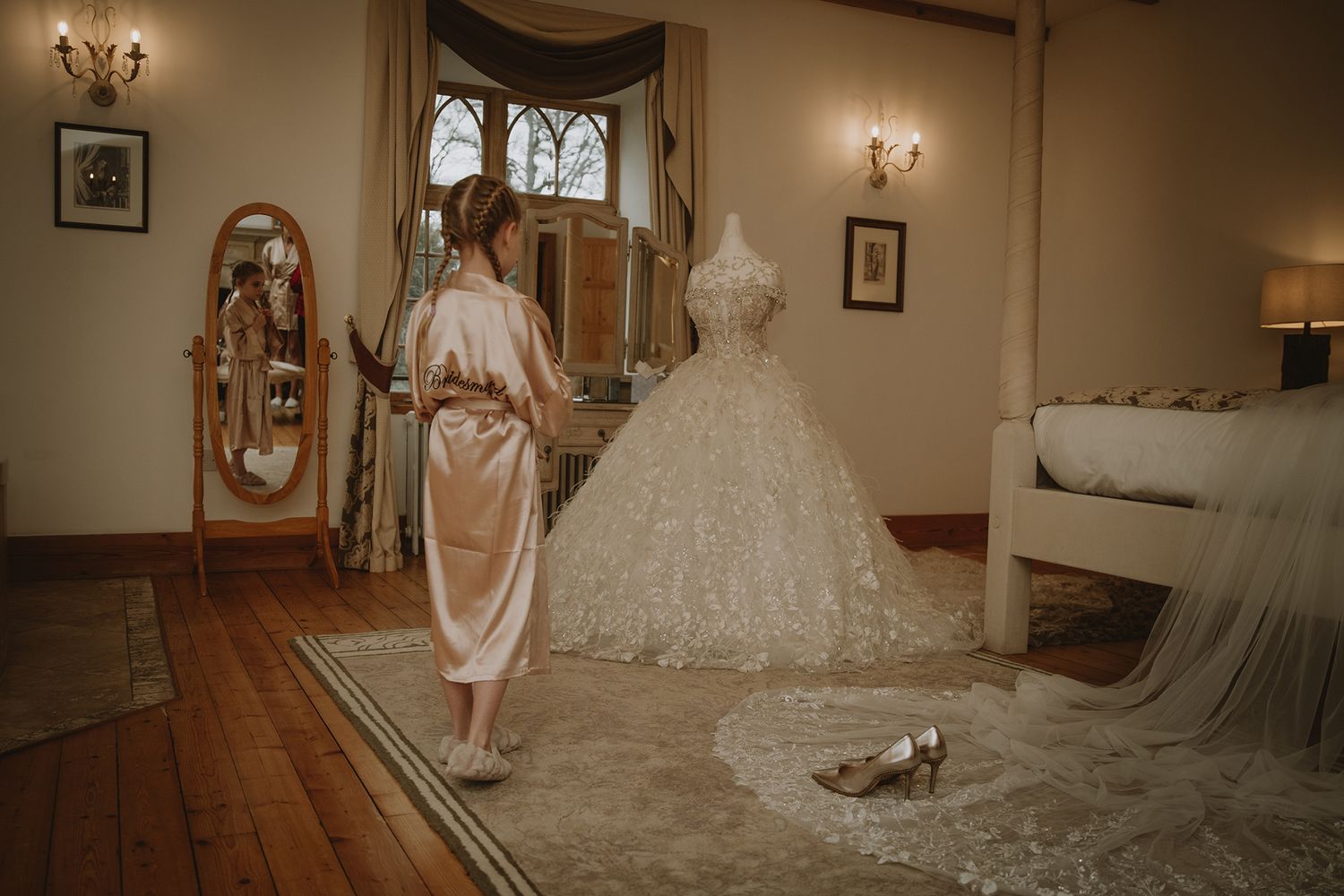 Bride in silk robe admiring elegant lace ball gown wedding dress on mannequin in luxurious bridal suite.