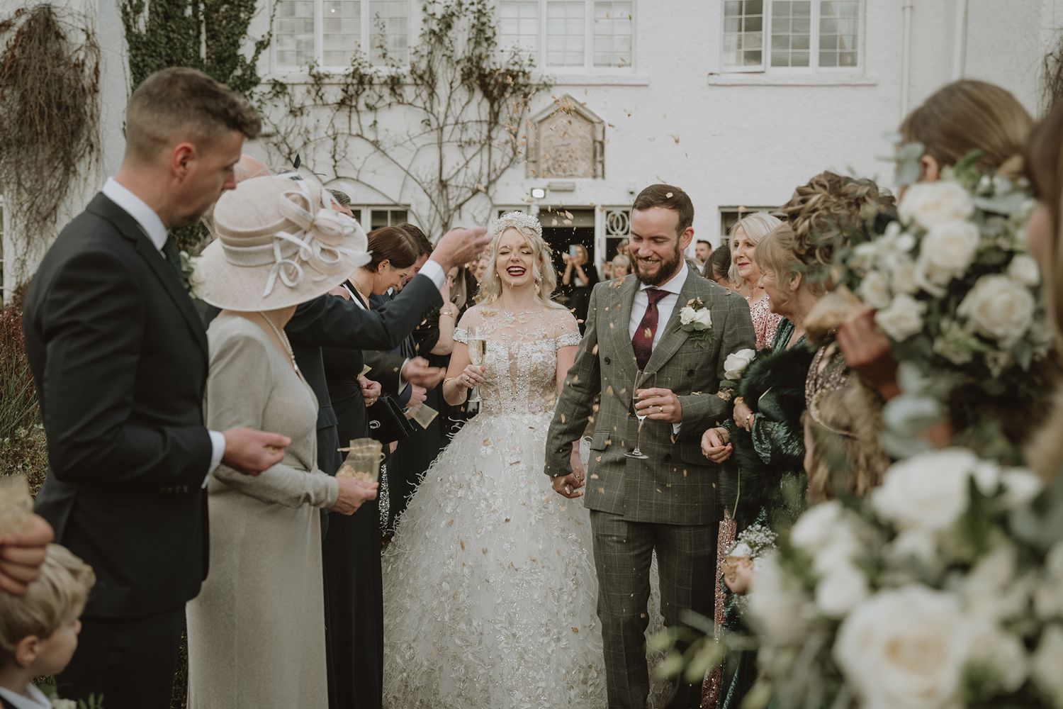 Bride and groom walk through confetti shower surrounded by smiling wedding guests outside elegant white venue.
