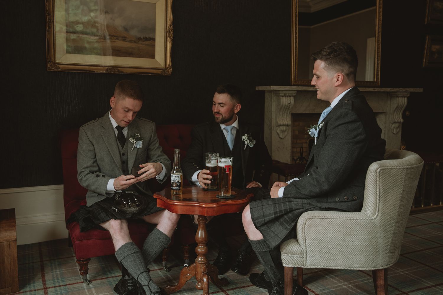 Three groomsmen in kilts and tweed suits relaxing with pints at pub before wedding.