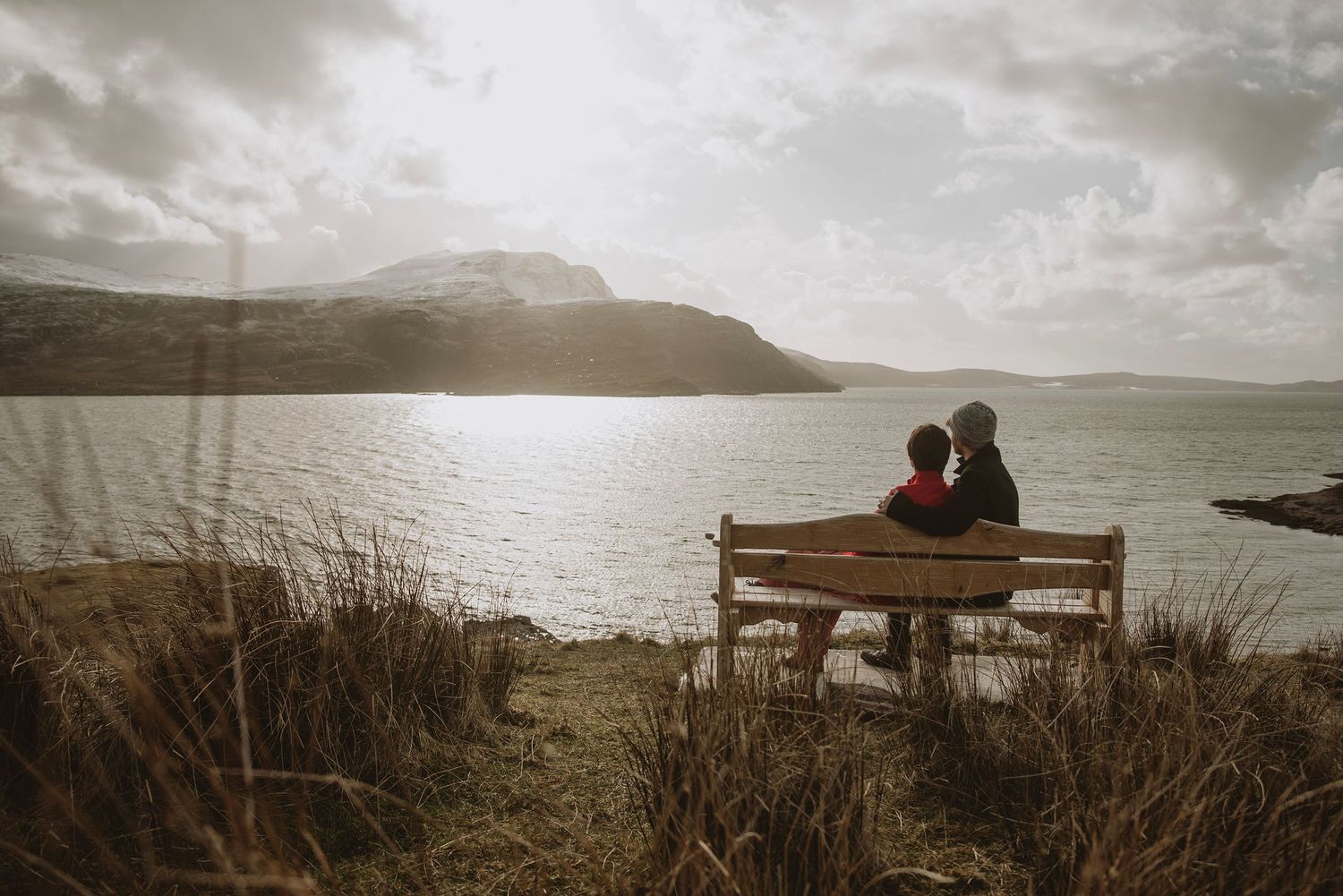 Couple sitting on a wooden bench overlooking a serene loch with misty mountains in the background.