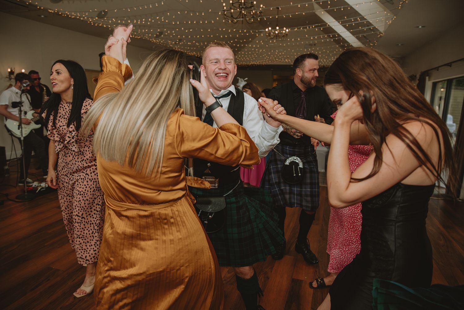 Joyful Scottish wedding guests dancing together, man in kilt laughing surrounded by women in gold and pink dresses.